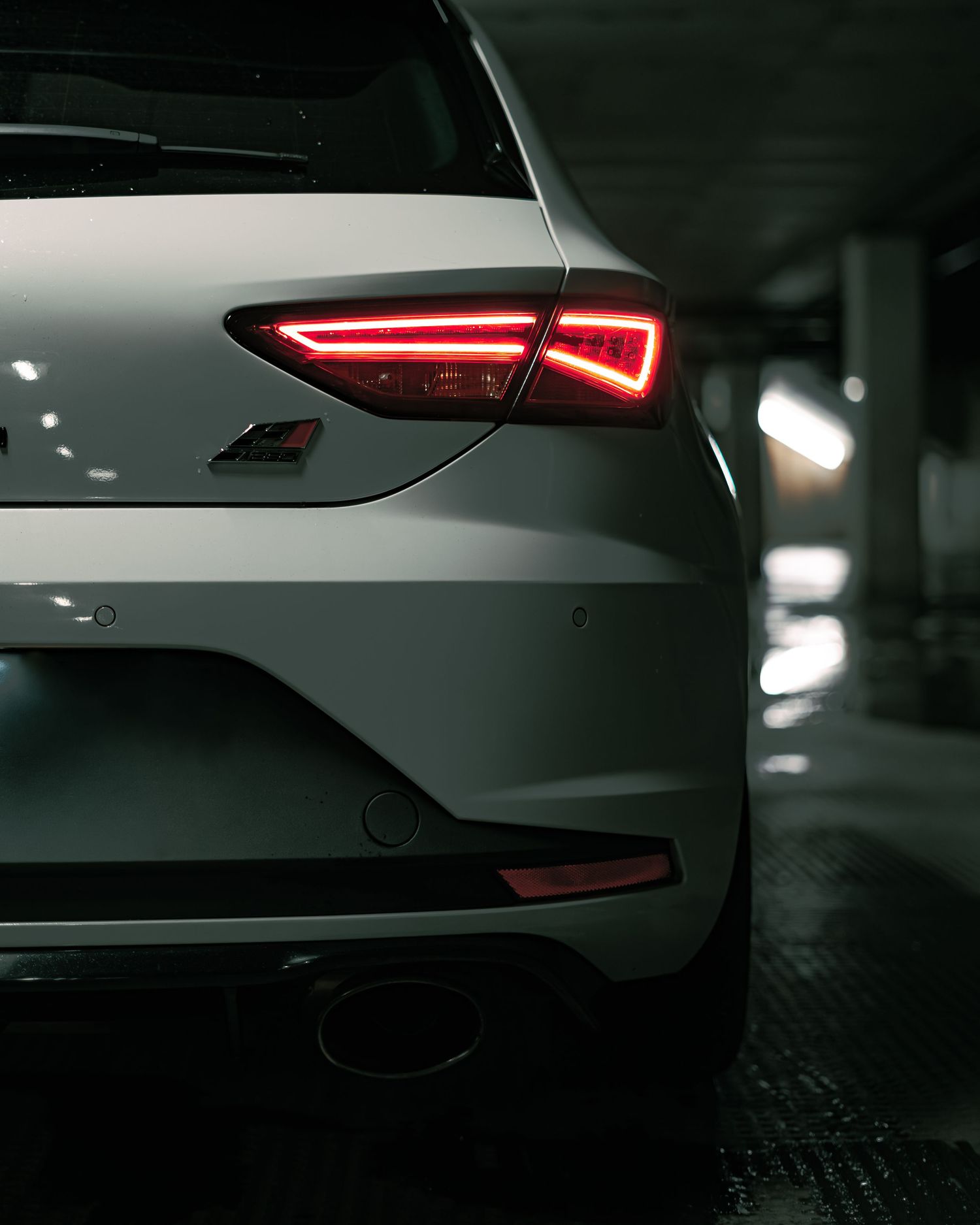 Rear view of white SEAT Leon Cupra showing glowing red LED tail lights in dark parking garage.