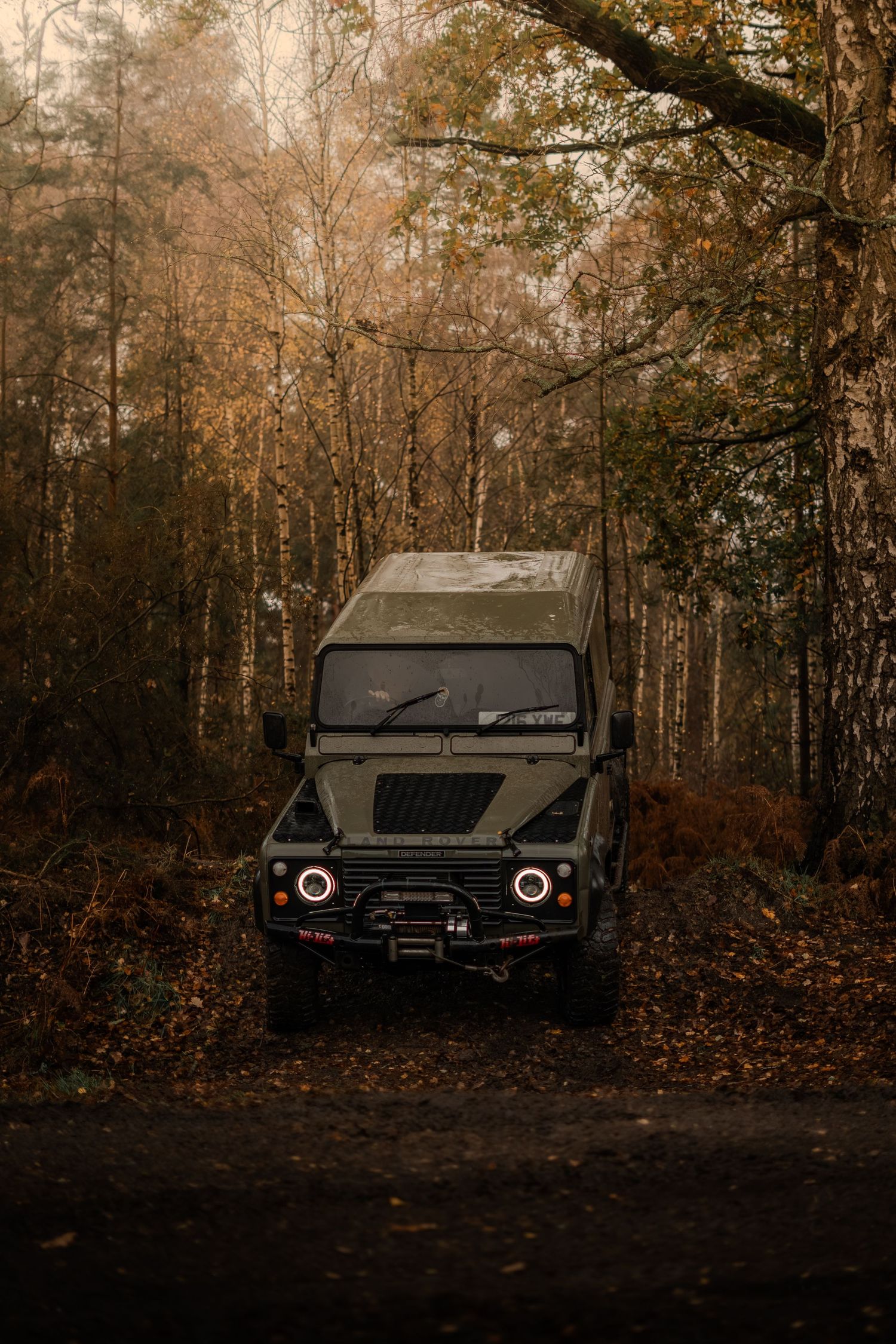 Dark green Land Rover Defender with halo headlights parked on a forest path surrounded by golden autumn trees.