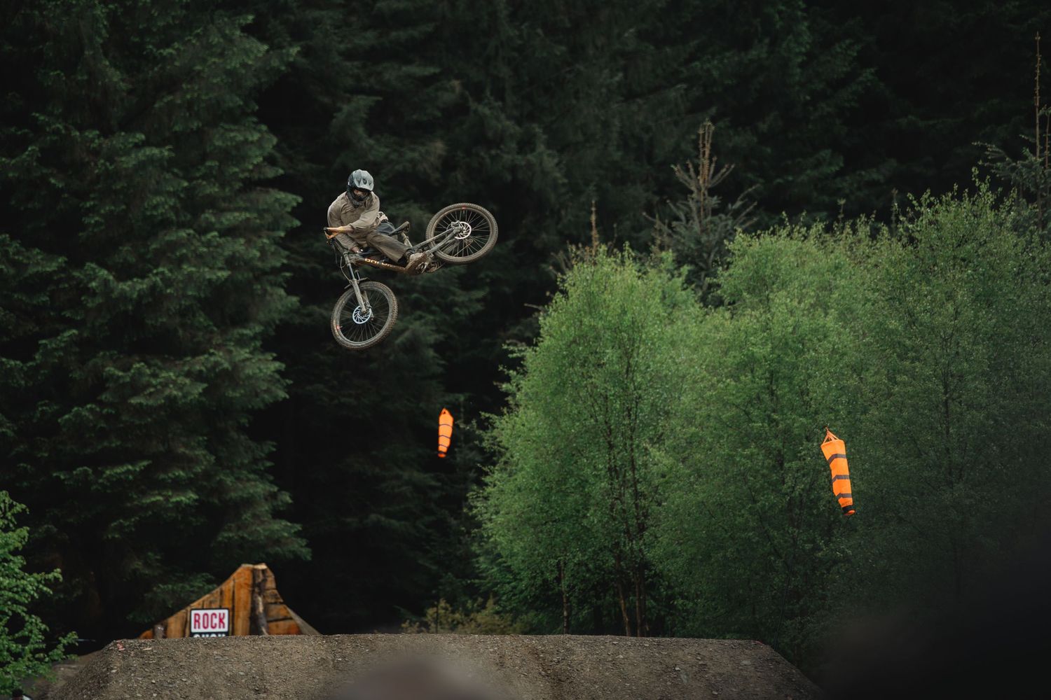 A mountain biker performs a high aerial jump trick over a dirt ramp surrounded by dense green forest trees.