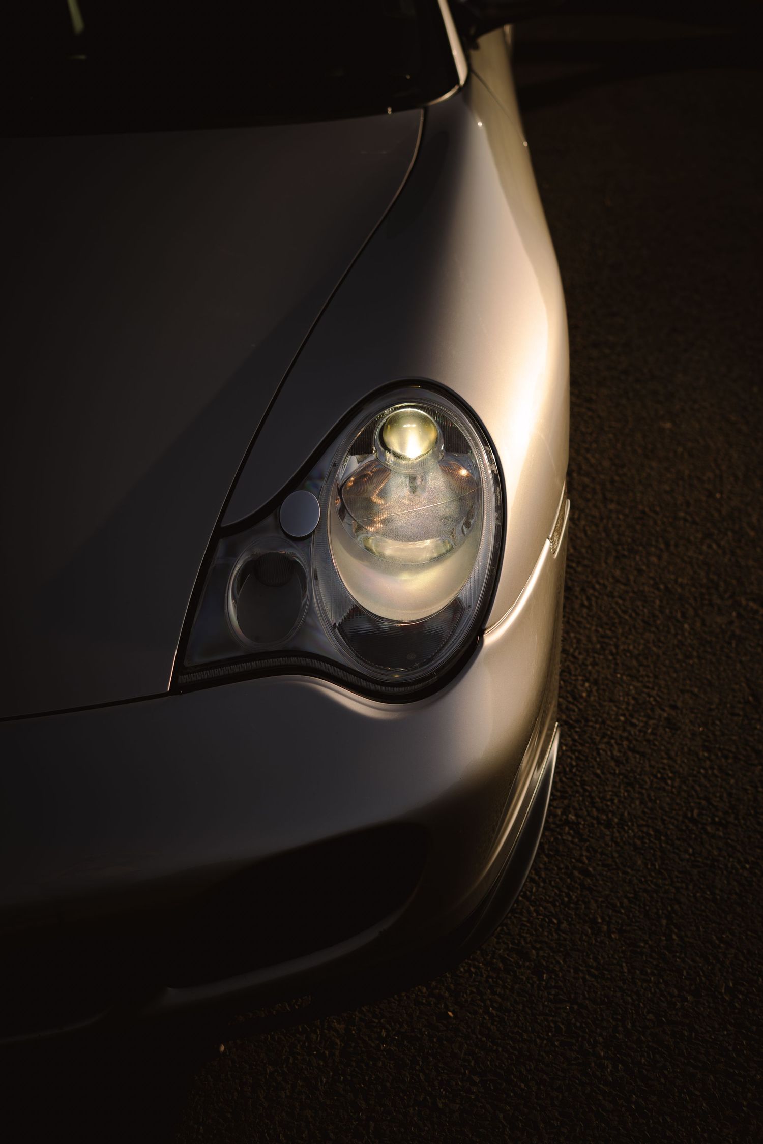 Close-up of a silver Porsche 911 headlight illuminated against a dark background on asphalt.