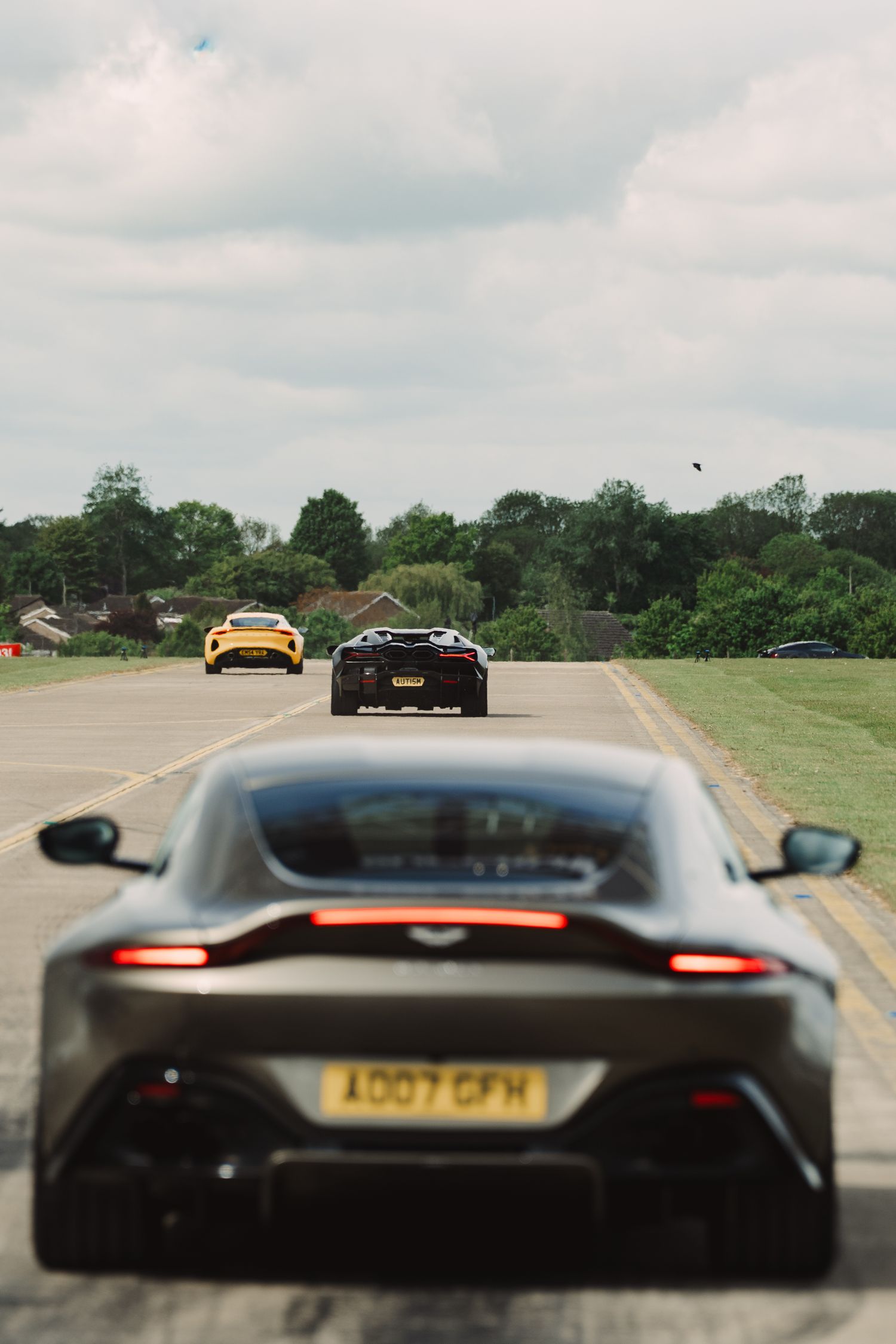 Three sports cars including an Aston Martin Vantage drive along a straight road surrounded by green trees under cloudy skies.