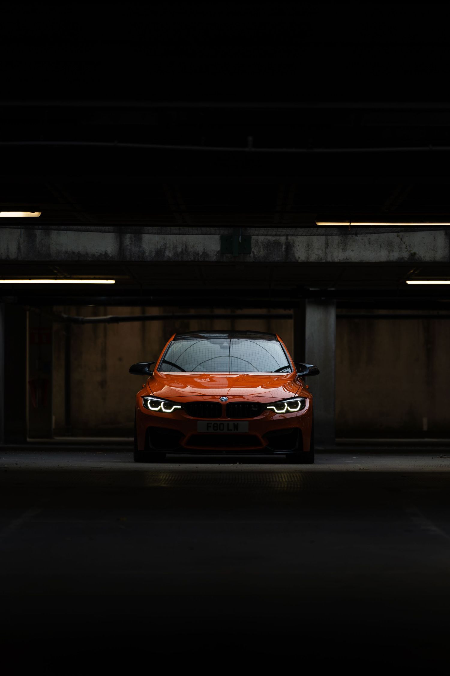 An orange BMW M4 parked in a dark underground garage, illuminated by overhead fluorescent lights.