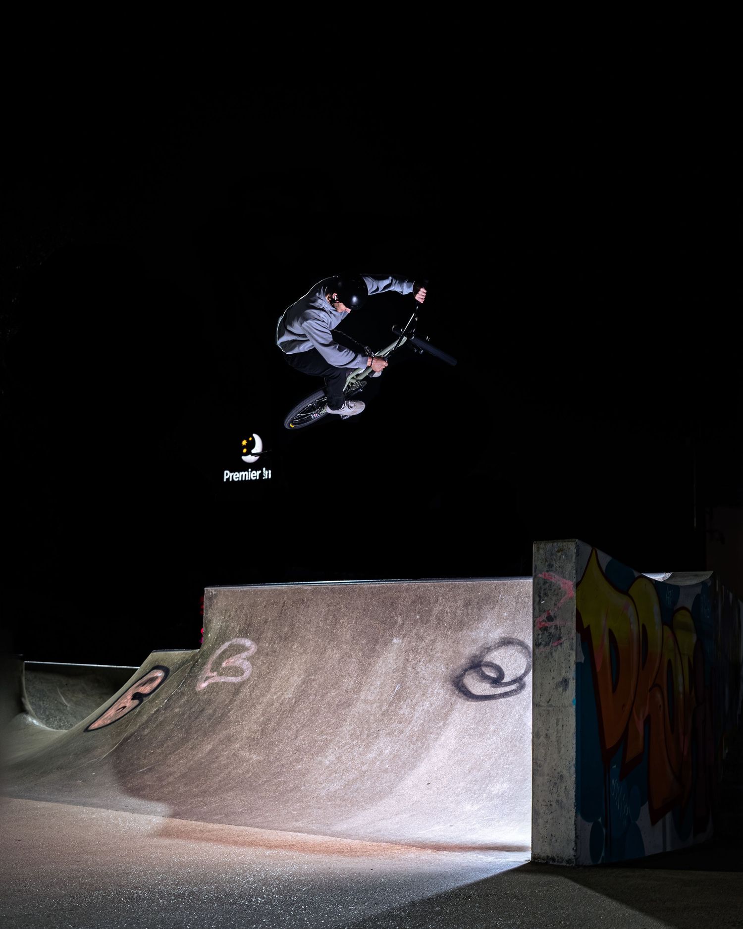BMX rider executing an impressive nighttime jump over a concrete skatepark ramp with graffiti walls.