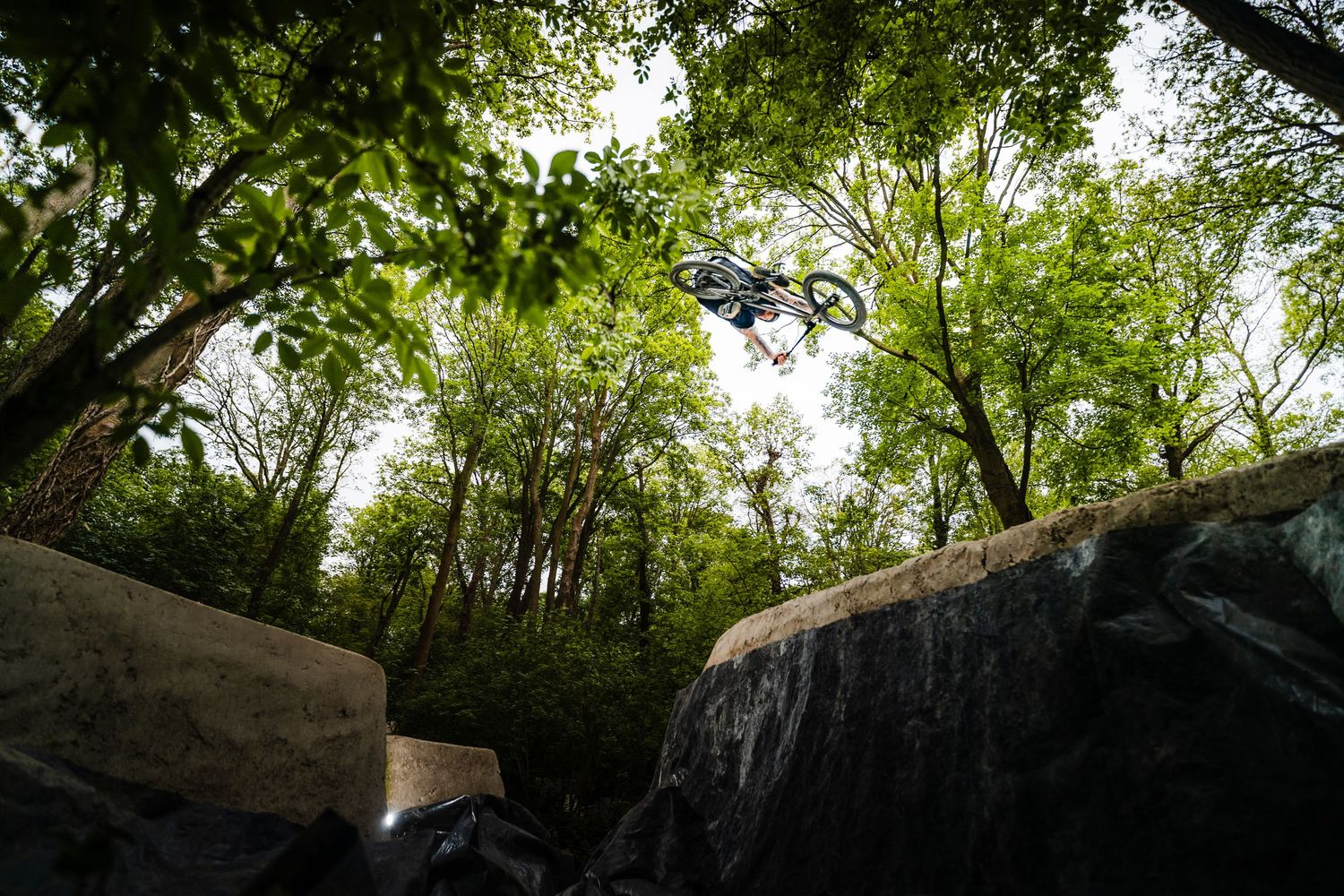 BMX rider performing a high aerial trick over rocky terrain surrounded by lush green trees outdoors.