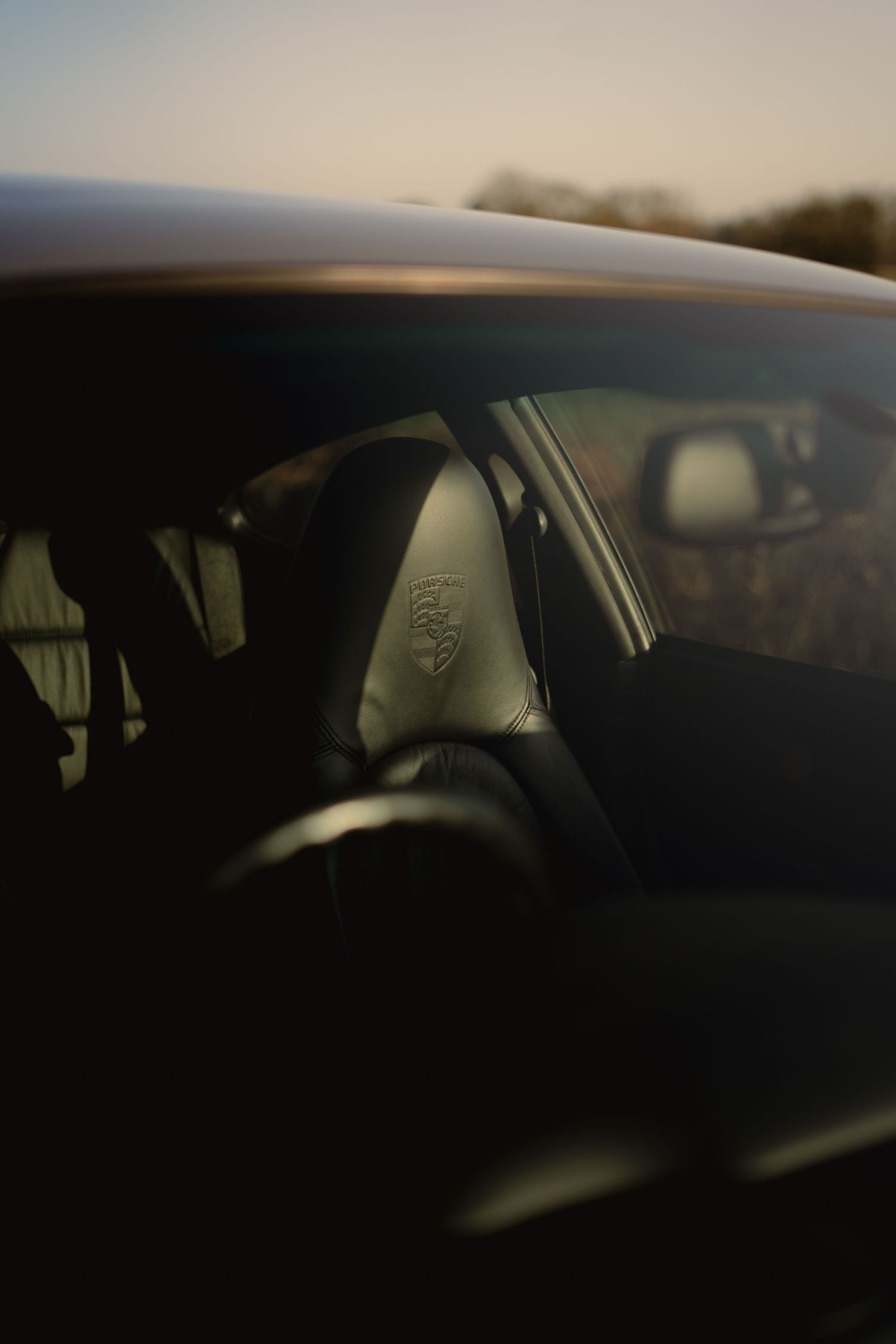Dark interior view through the window of a Porsche 911 showing leather headrest with embossed logo at sunset.