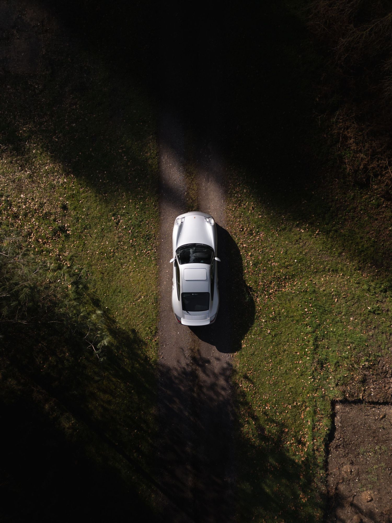 Overhead drone view of a white Porsche 911 on a narrow gravel path with grass verges in dappled sunlight.