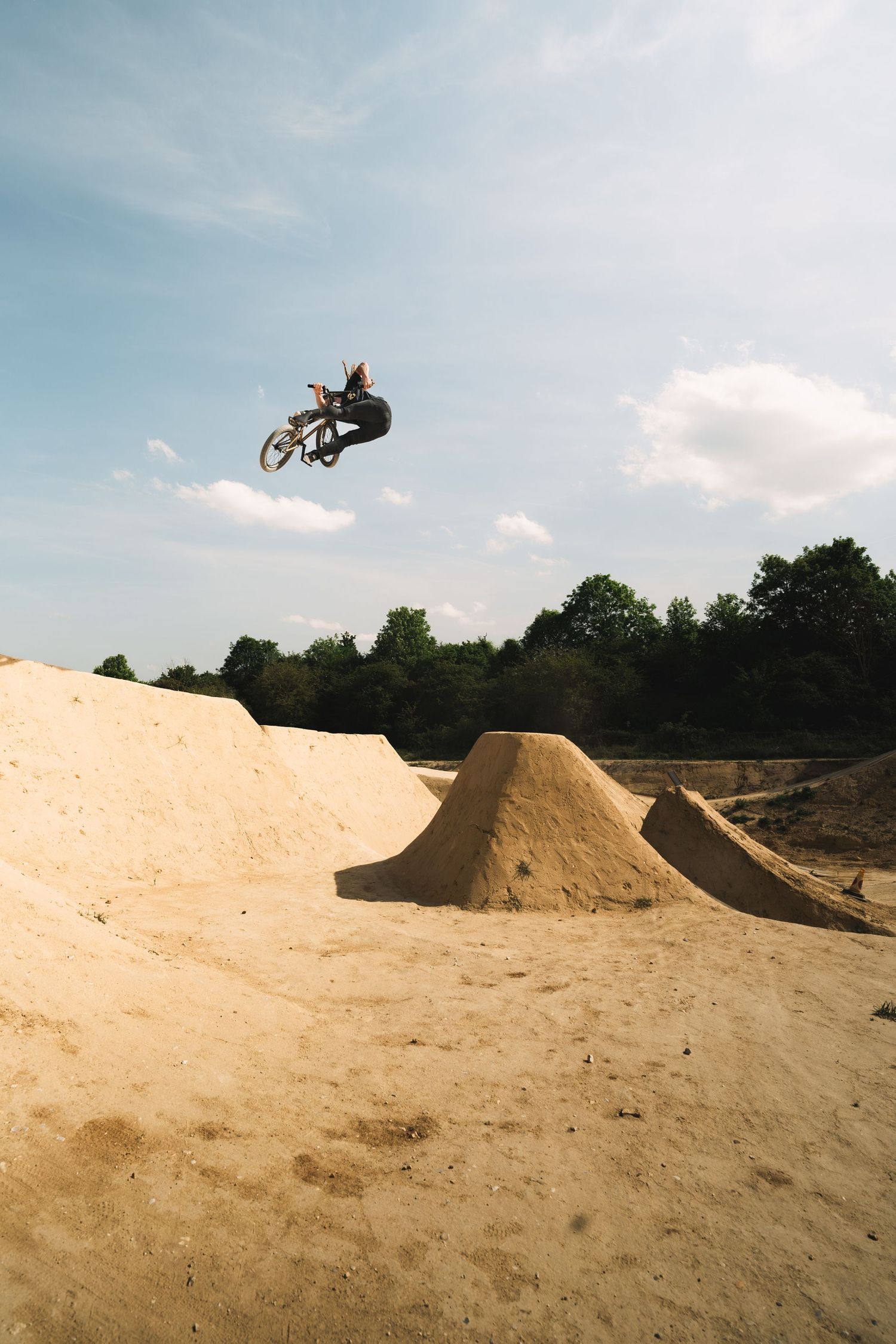 BMX rider soaring high above sandy dirt jumps while performing a trick at an outdoor bike park under a cloudy sky.