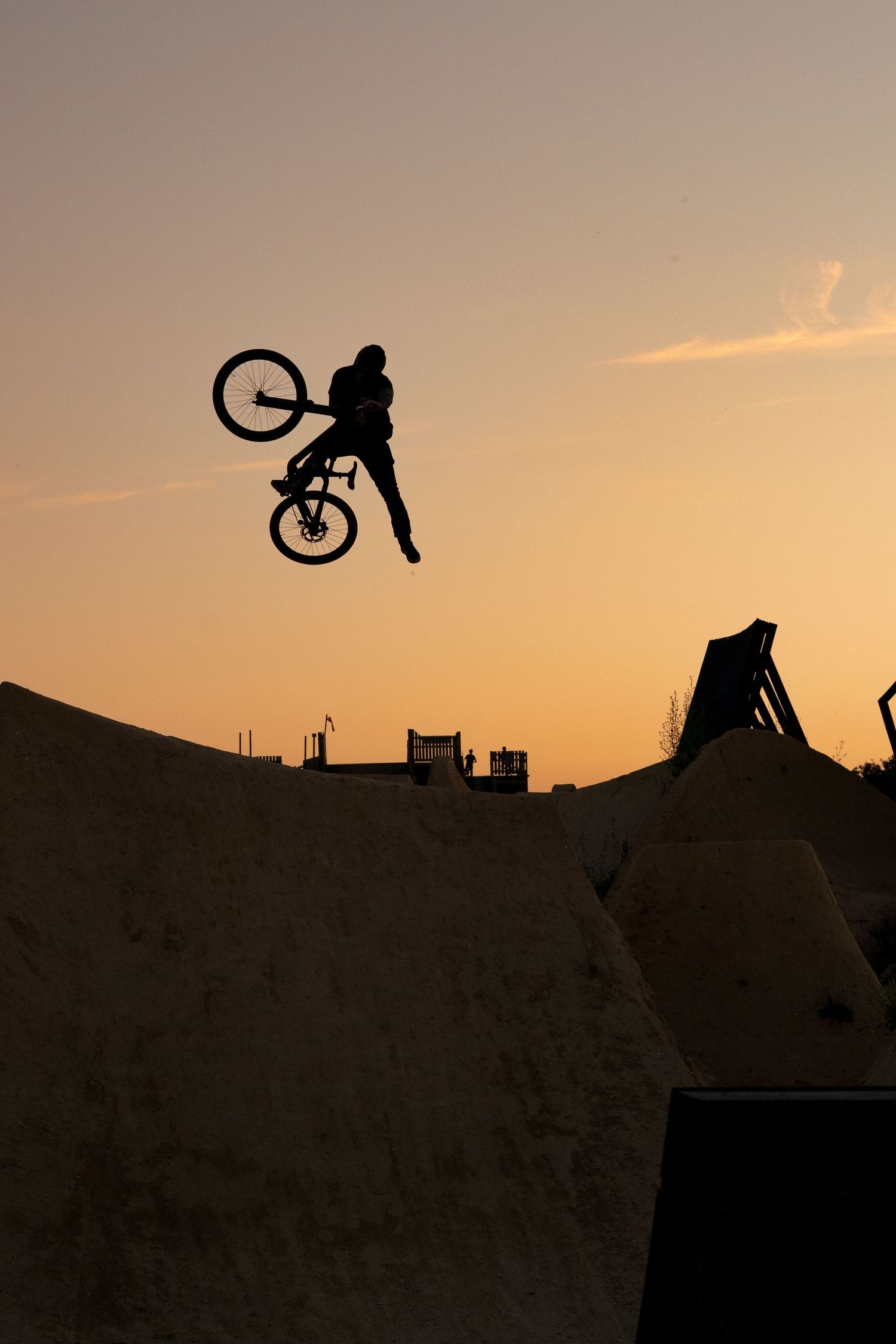 Silhouette of a BMX rider performing a high jump trick over a skate park ramp at sunset with orange sky background.