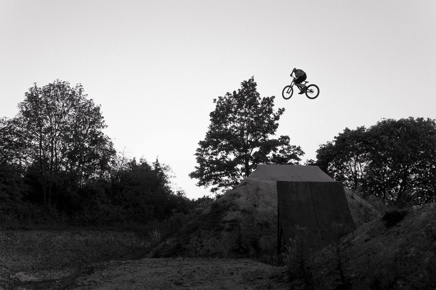 Black and white photo of a cyclist performing a high jump trick off a dirt ramp surrounded by trees.