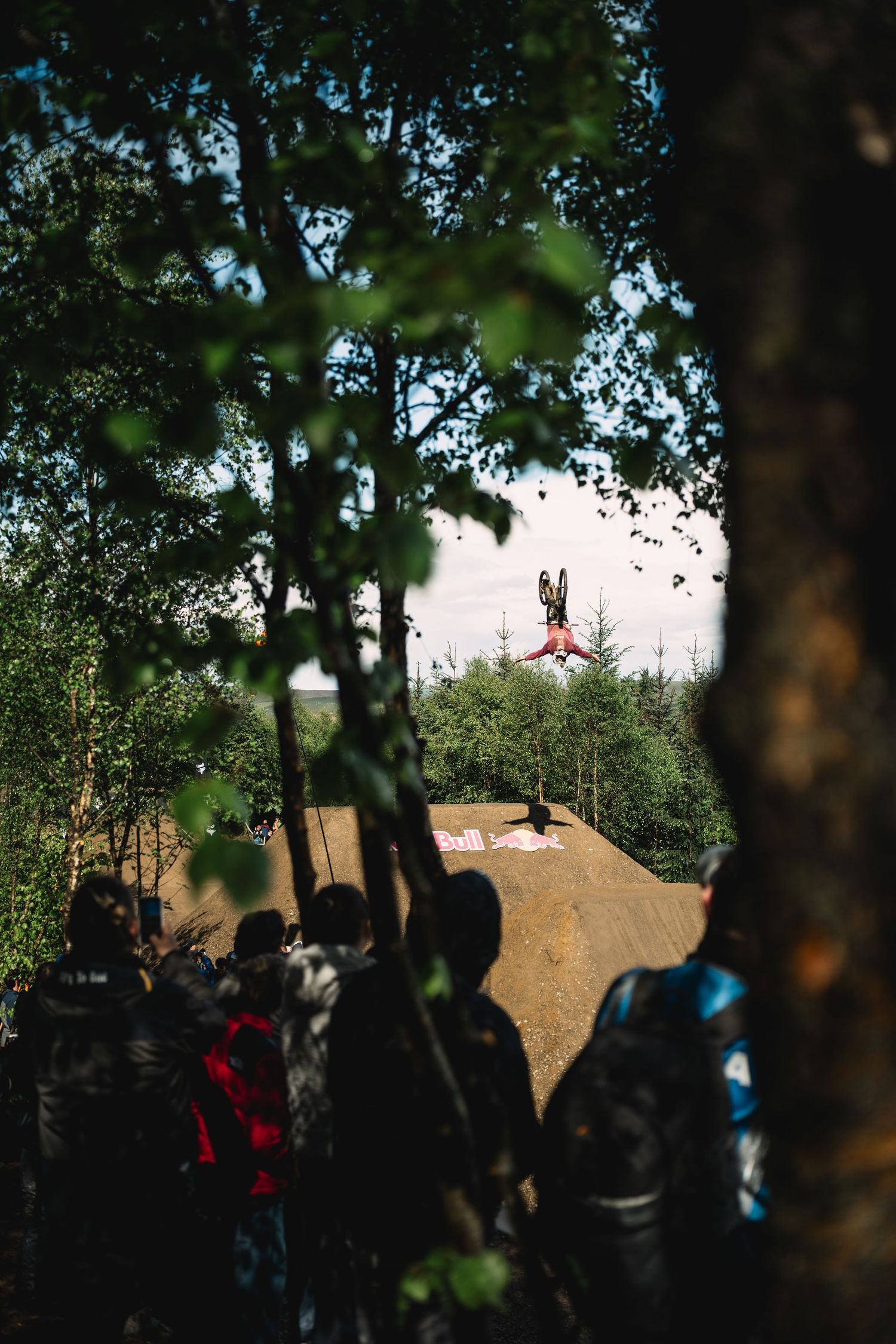 A BMX rider performs an aerial trick at a Red Bull event while spectators watch from behind trees.