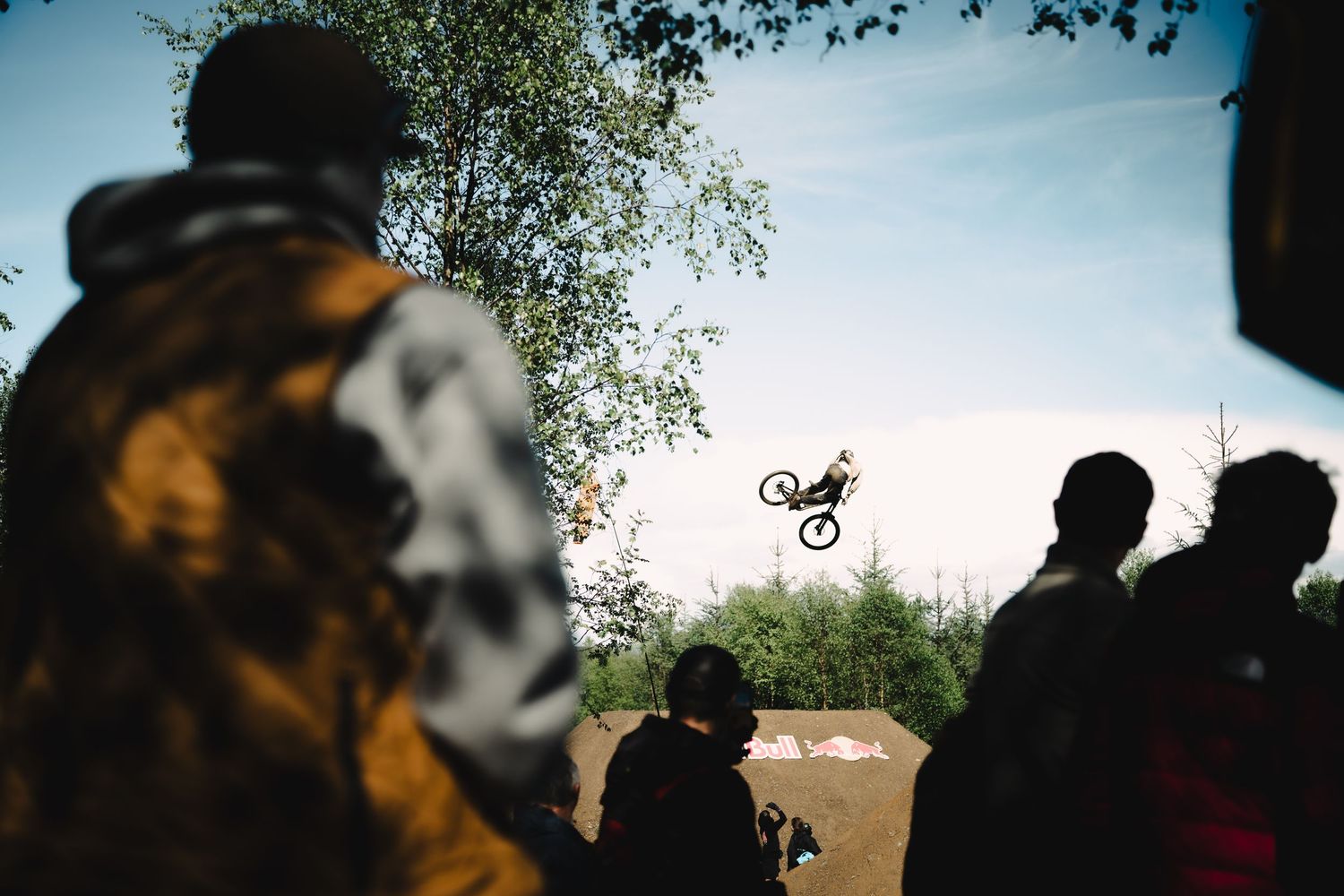 A BMX rider performs a high aerial trick while spectators watch from the ground at an outdoor dirt jump park.