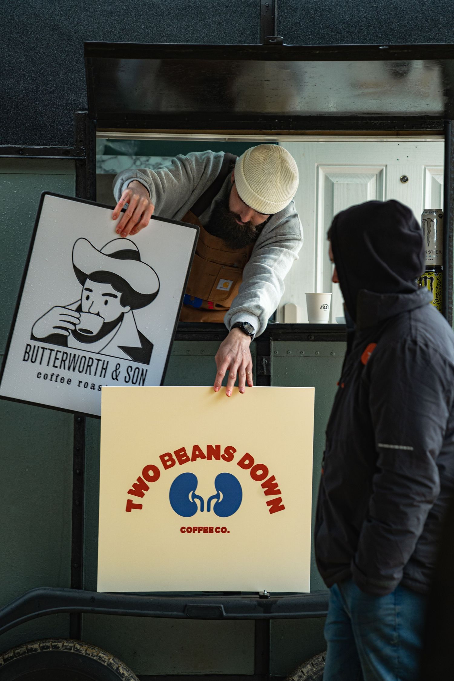 Bearded barista in white beanie passes Two Beans Down Coffee Co. sign from mobile coffee trailer to customer.