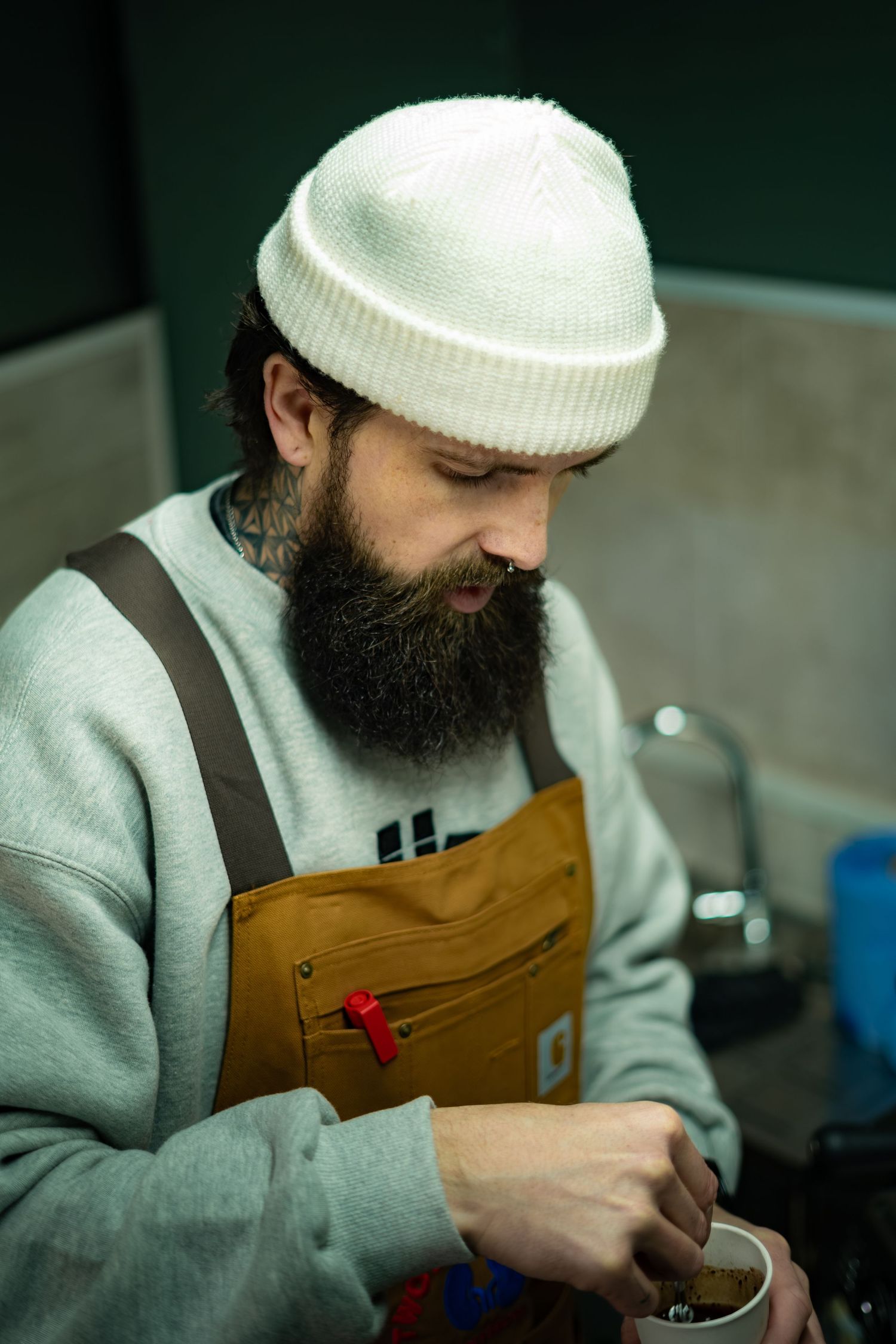 Focused bearded barista in white beanie and brown apron prepares coffee grounds in small cup.