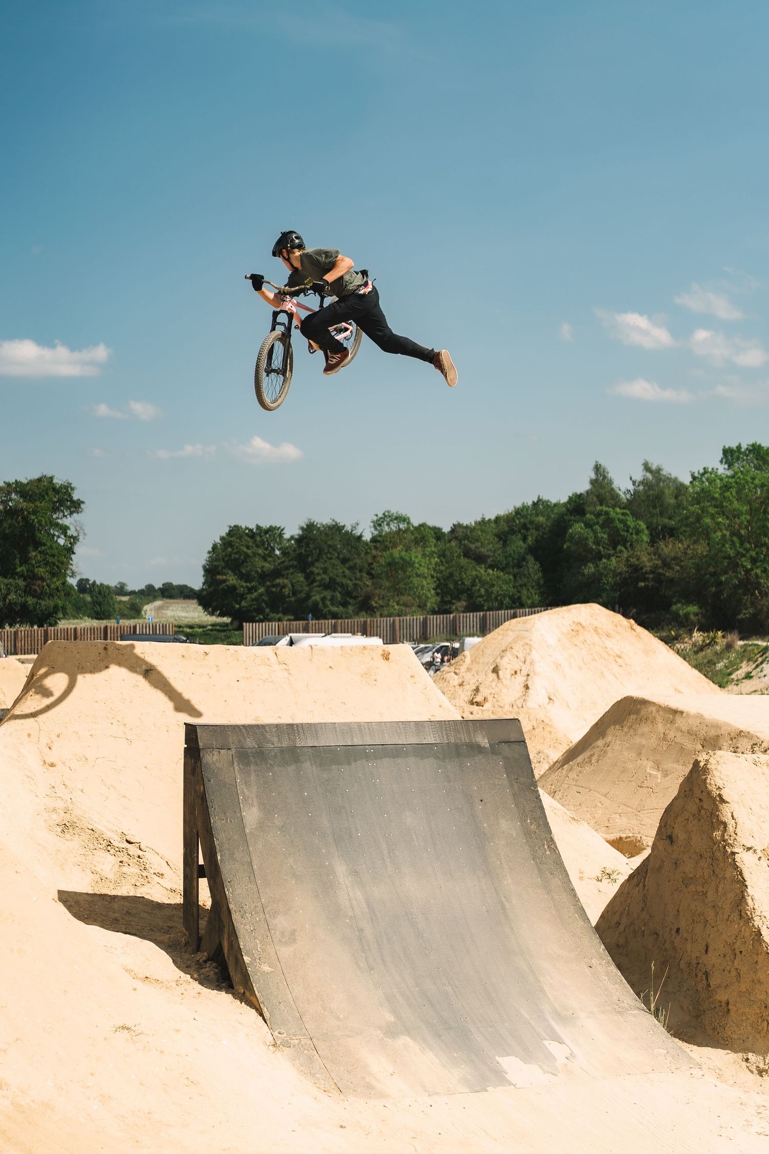 BMX rider performing a high aerial trick off a dirt ramp at an outdoor bike park under a clear blue sky.