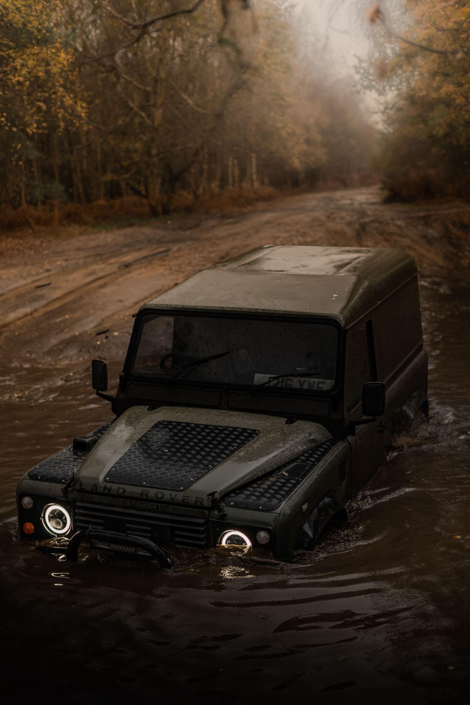 Land Rover Defender wading through a deep muddy water crossing on a foggy autumn forest trail.