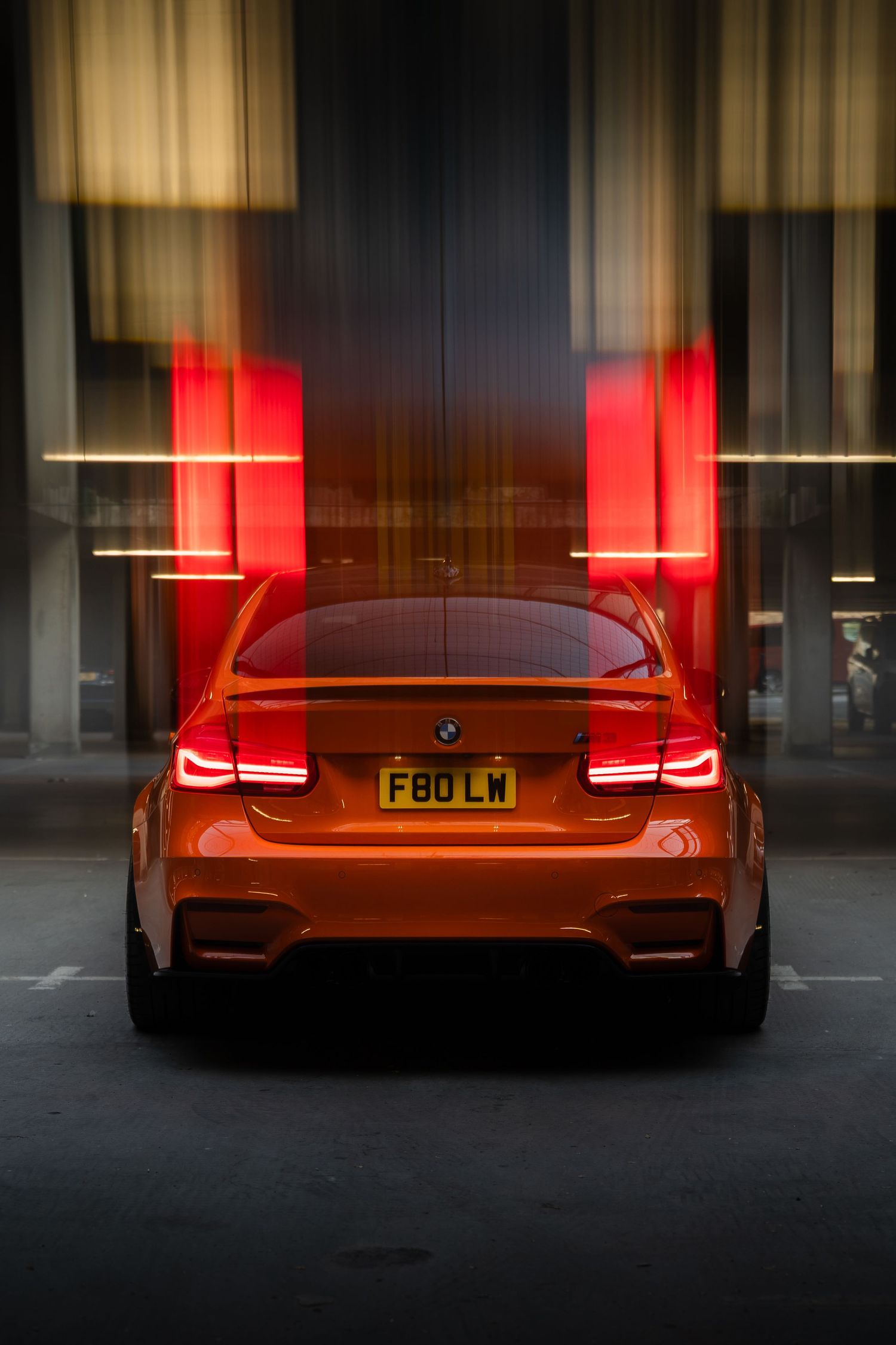 Rear view of an orange BMW M3 F80 LM with glowing taillights, captured with motion blur effect at night.