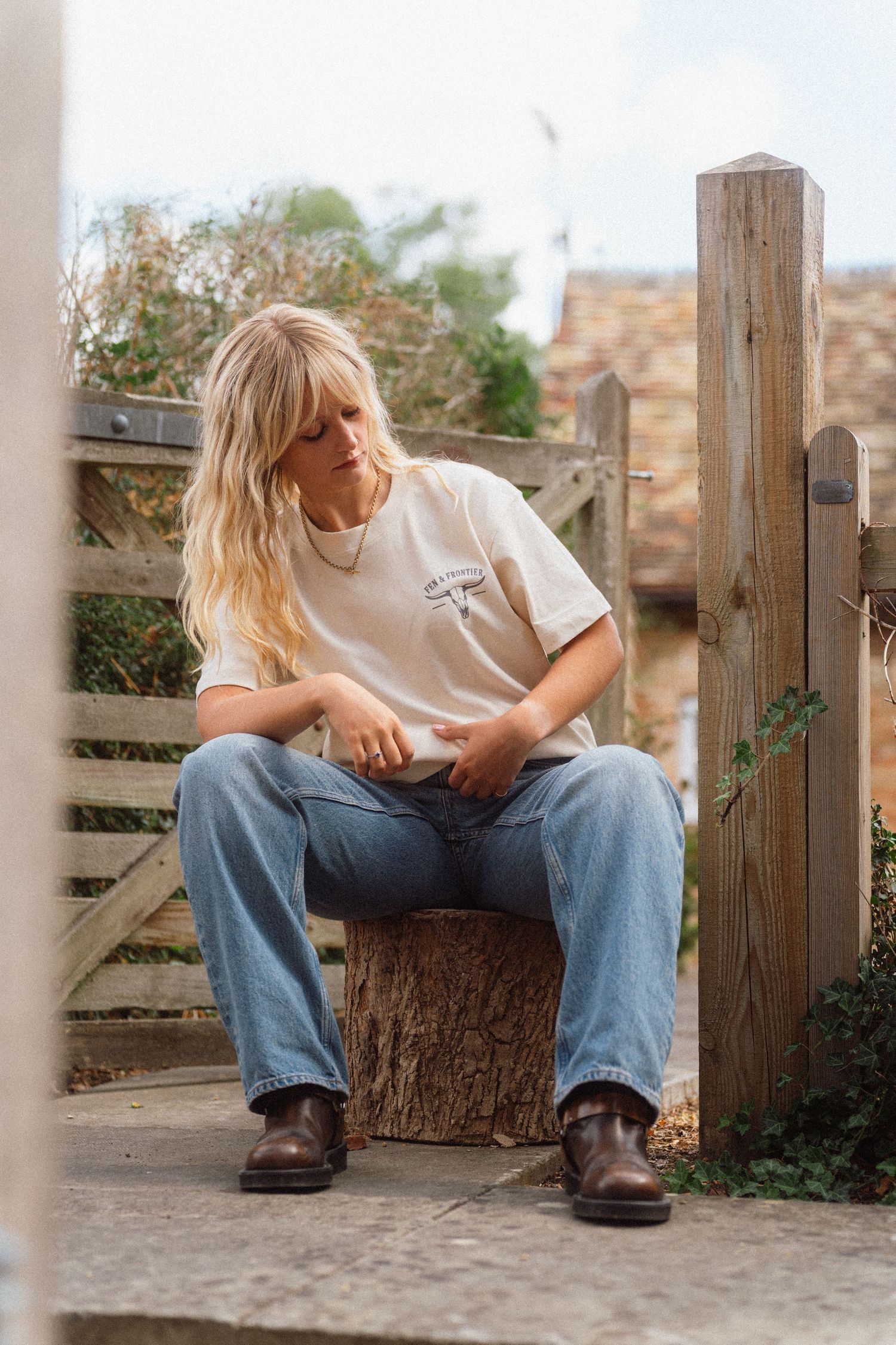 Blonde woman sitting on a tree stump by a wooden fence, wearing a white tee, blue jeans, and brown boots.
