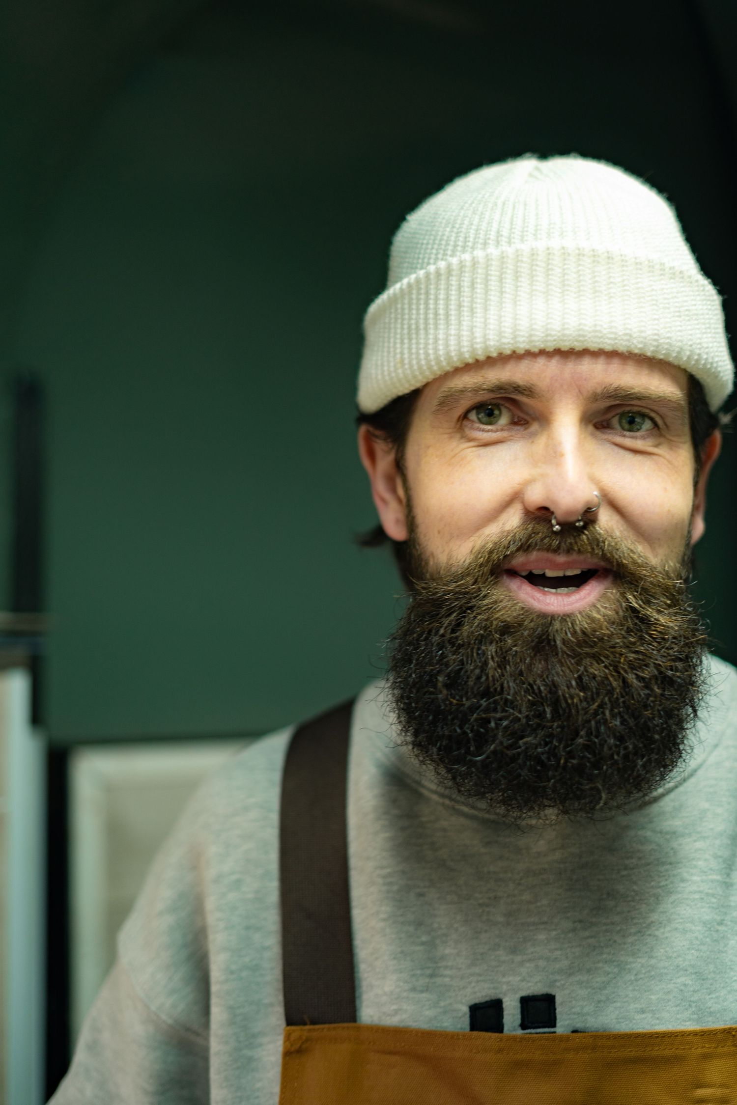 A bearded barista wearing a white beanie hat and brown apron smiles in a coffee shop.