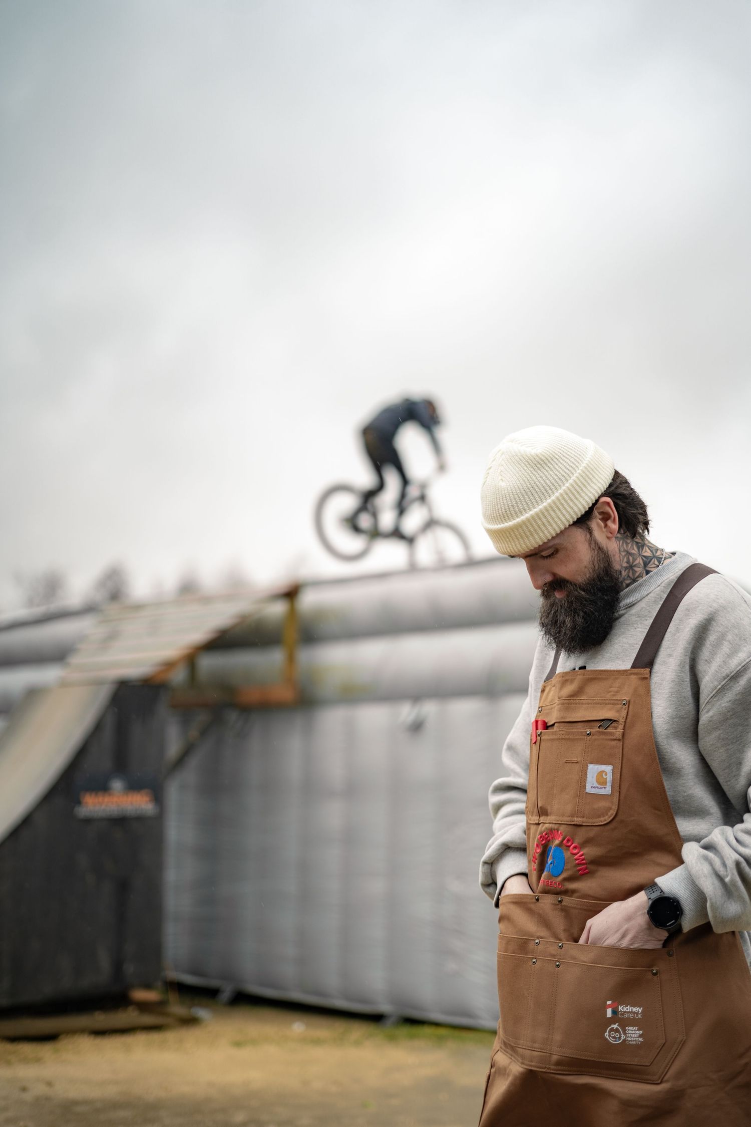 A bearded barista in a brown Carhartt apron watches a BMX rider on a ramp at an outdoor park.