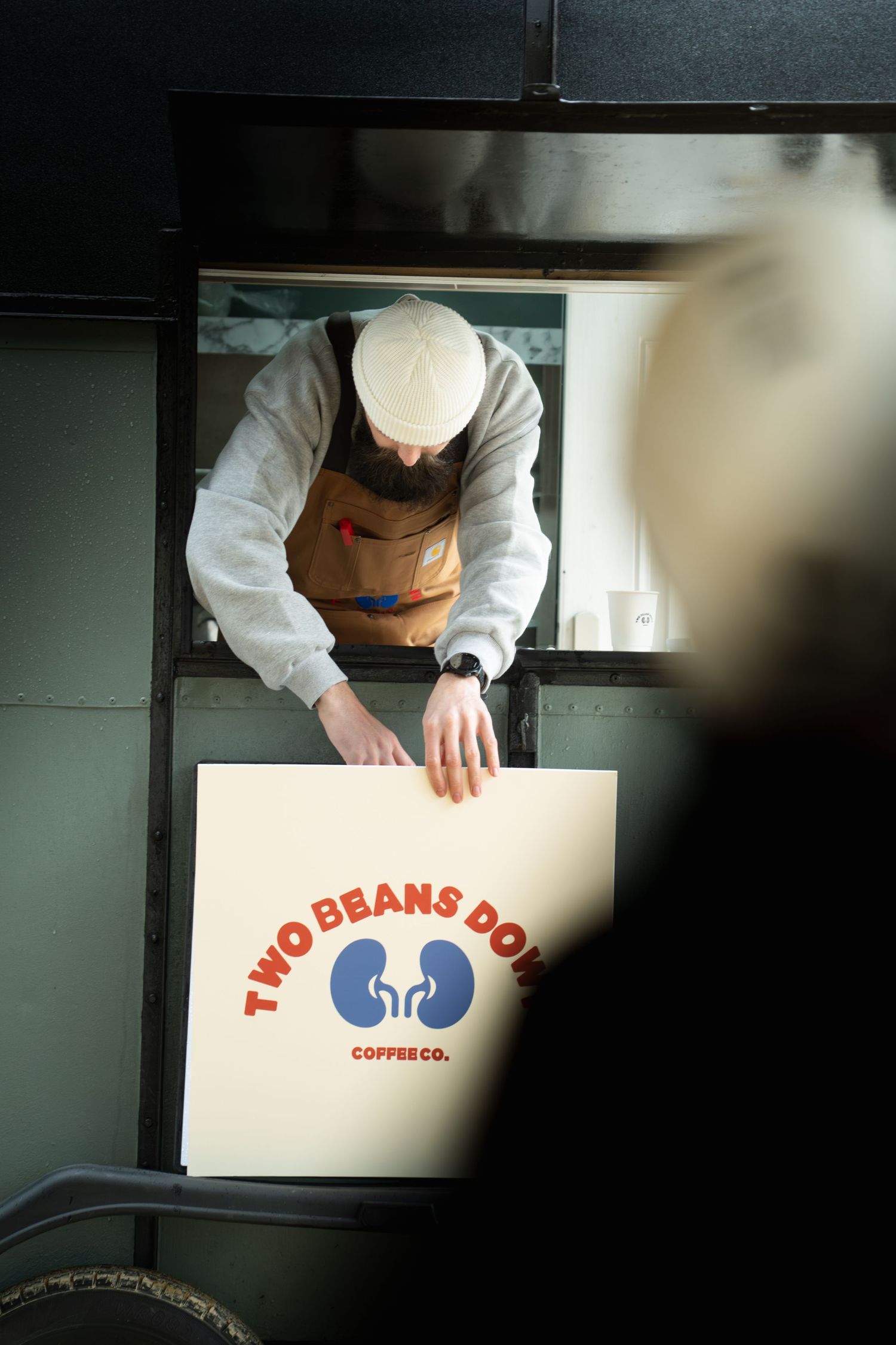 Overhead view of bearded barista in white beanie handling Two Beans Down Coffee Co. signage in mobile cafe.