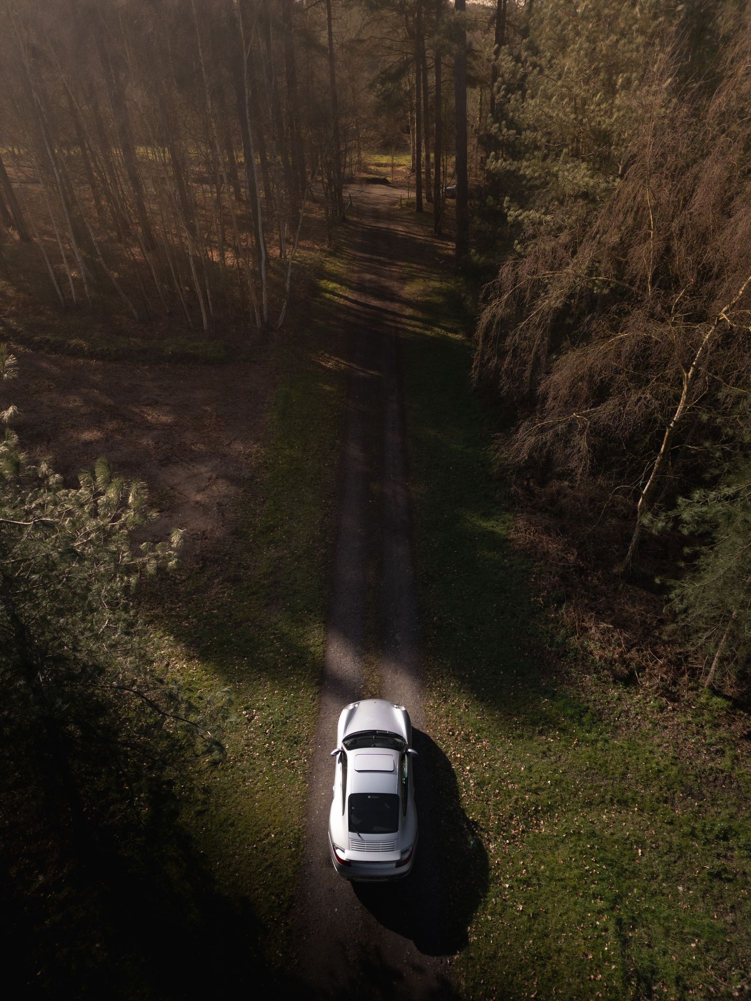 Drone aerial view of a silver Porsche 911 parked on a shaded forest dirt road flanked by tall trees.