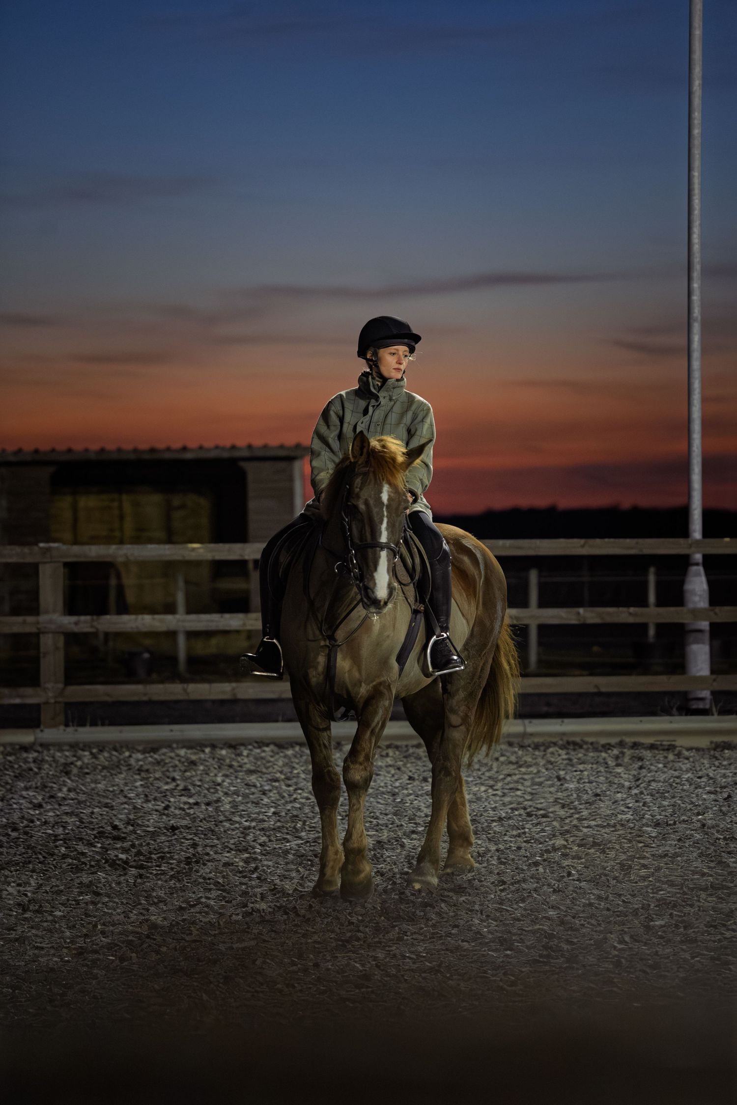 Young rider on horseback at dusk in an outdoor equestrian arena with a vibrant sunset sky.