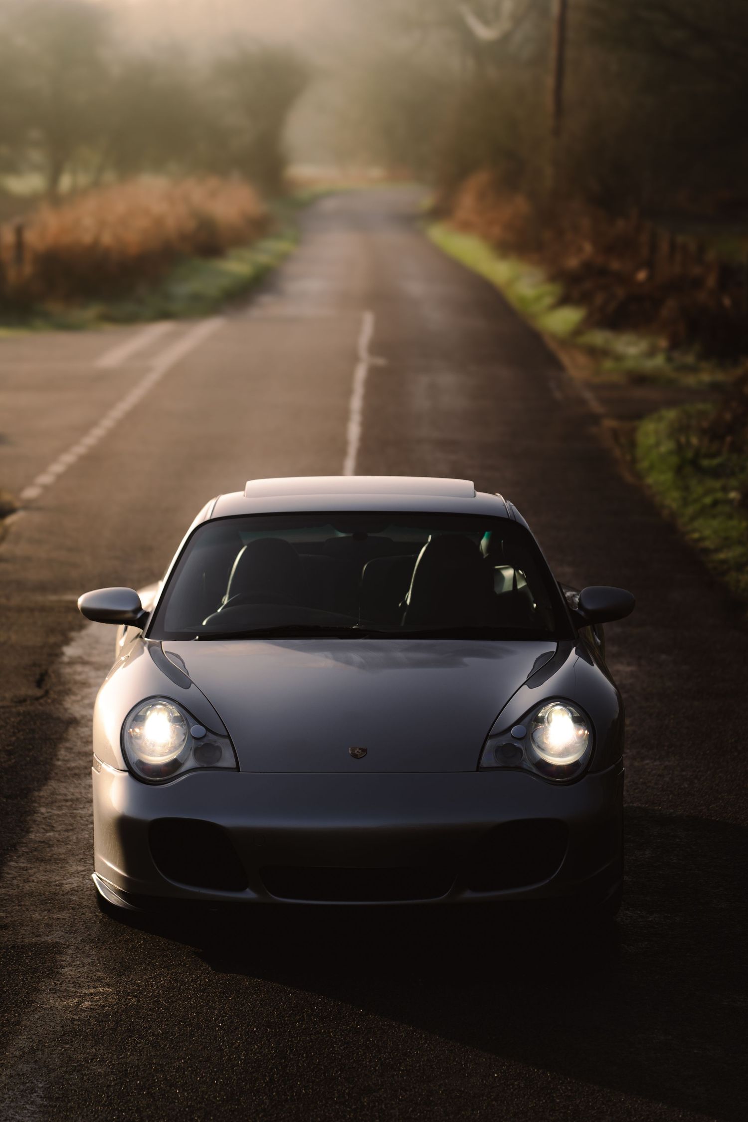 Silver Porsche 911 Turbo with headlights on facing camera on a foggy countryside road.