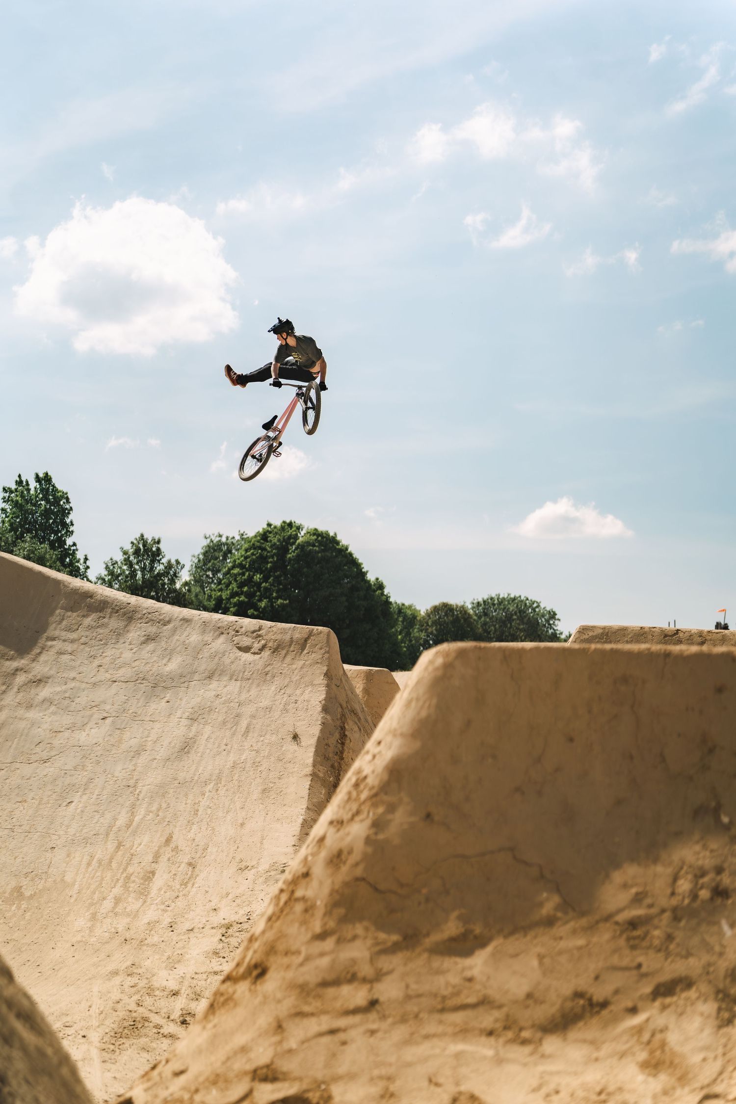 BMX rider performing a high jump trick off a dirt ramp at an outdoor bike park under a partly cloudy sky.