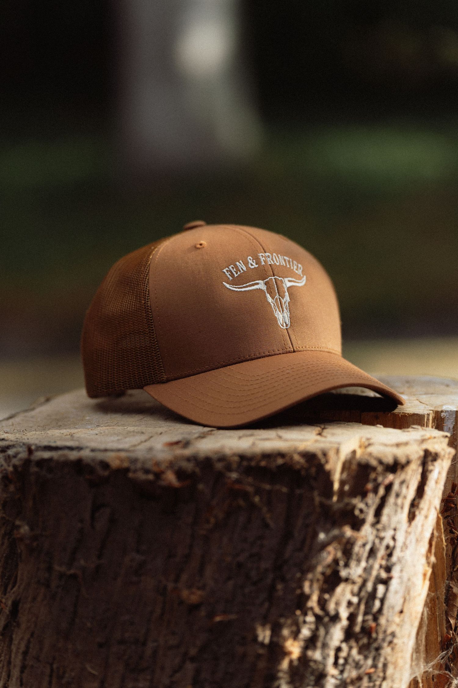 Brown trucker cap with white longhorn skull embroidery from Fen & Frontiers, displayed on a rustic wood stump outdoors.
