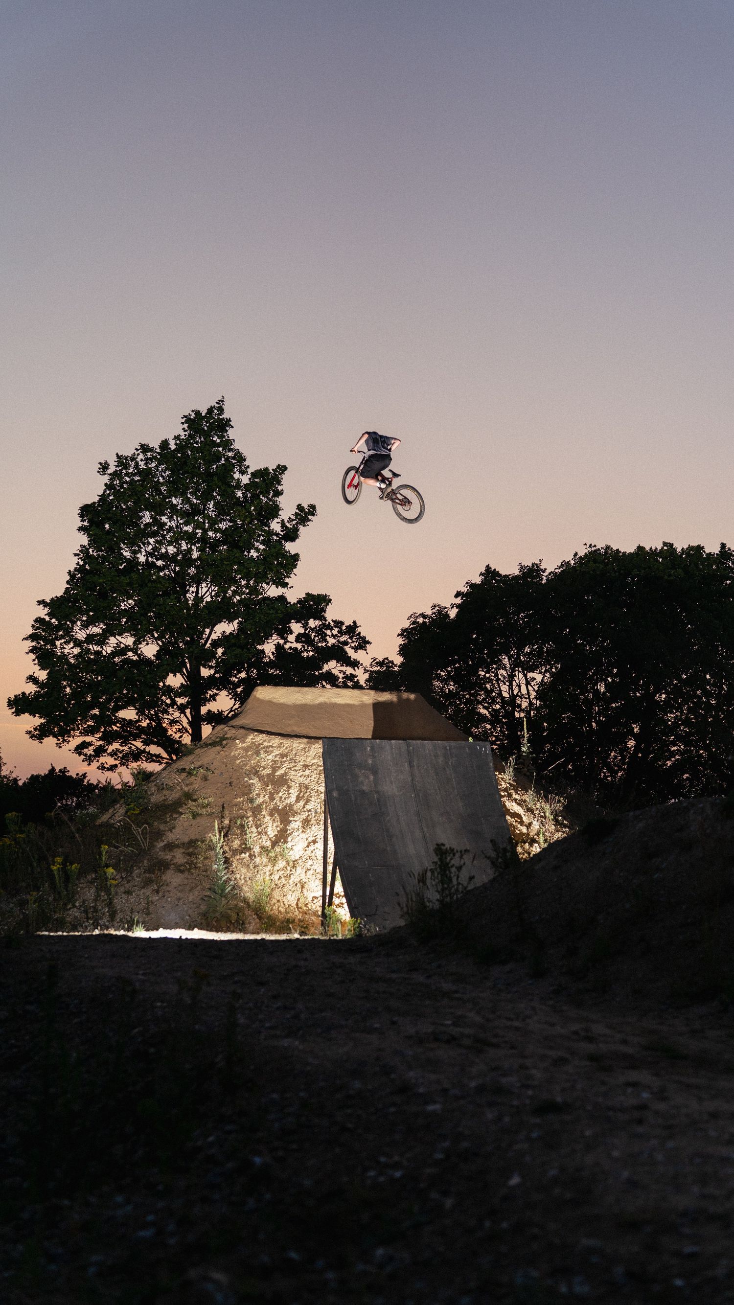 BMX rider performs a high aerial jump off a dirt ramp at dusk, silhouetted against a twilight sky with trees.
