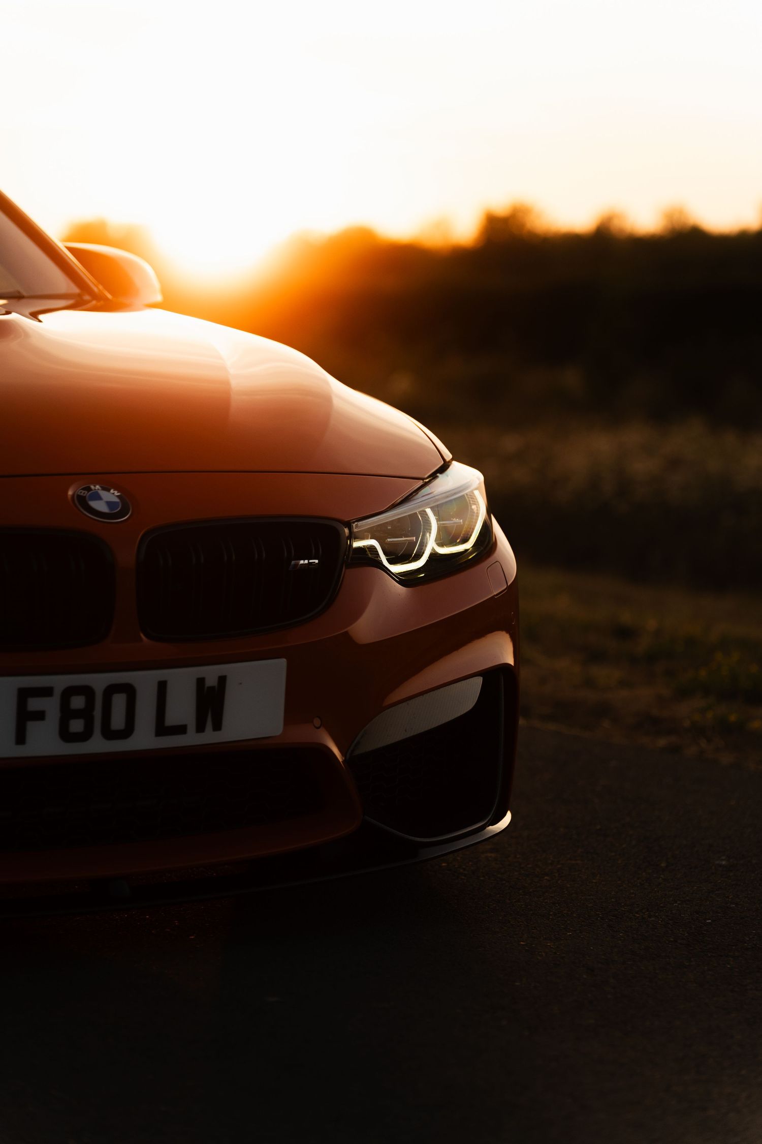 Orange BMW M4 F80 photographed at golden hour sunset on a country road, showcasing LED headlights and iconic kidney grille.