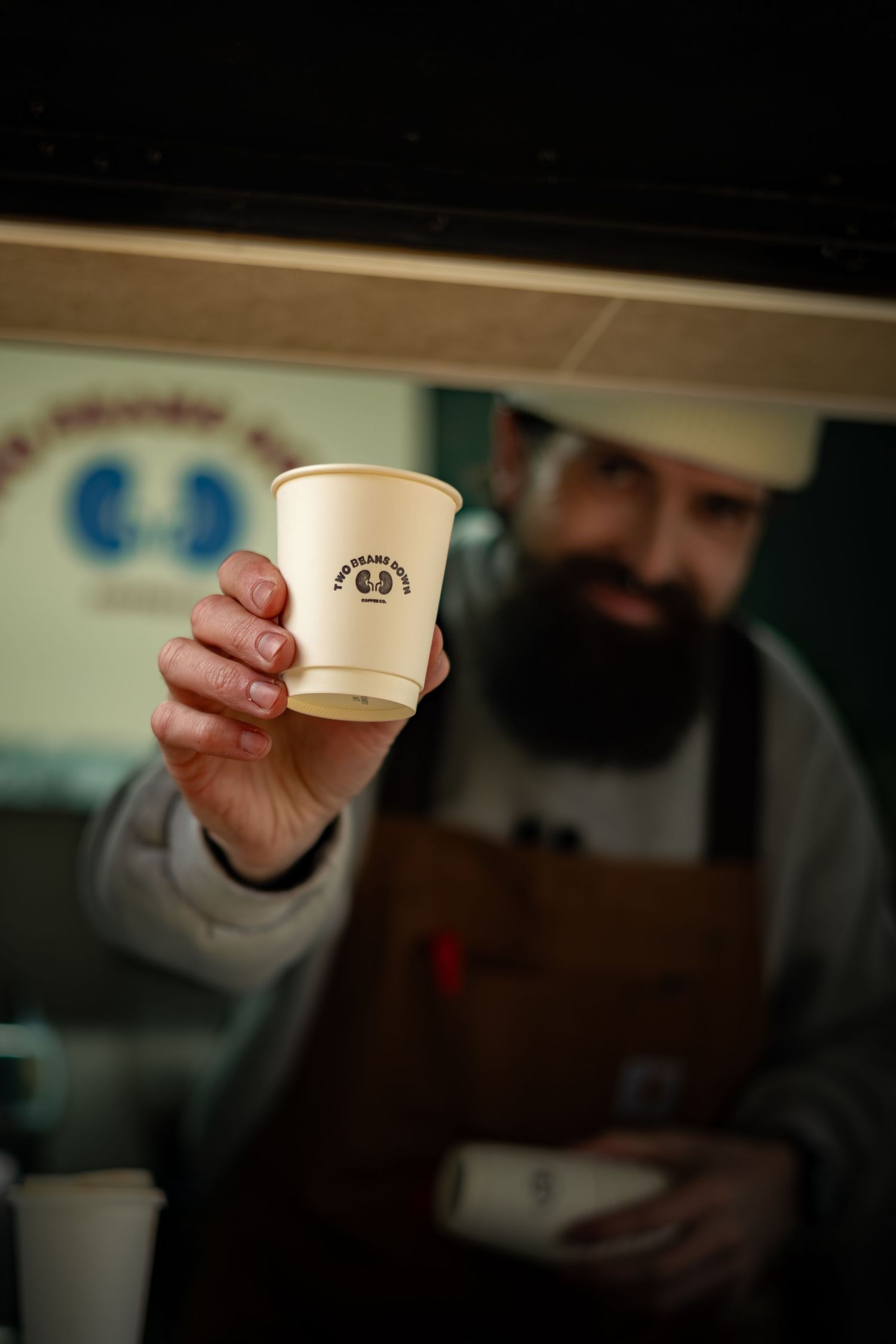 Bearded barista extends a branded paper coffee cup toward camera from a food truck service window.