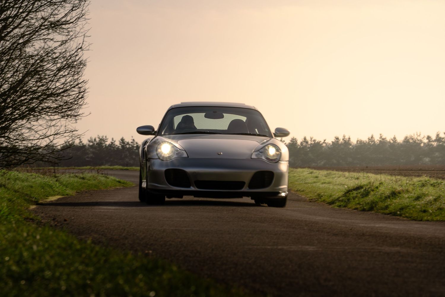 Silver Porsche 911 Turbo front view on a narrow country road with warm golden hour lighting.