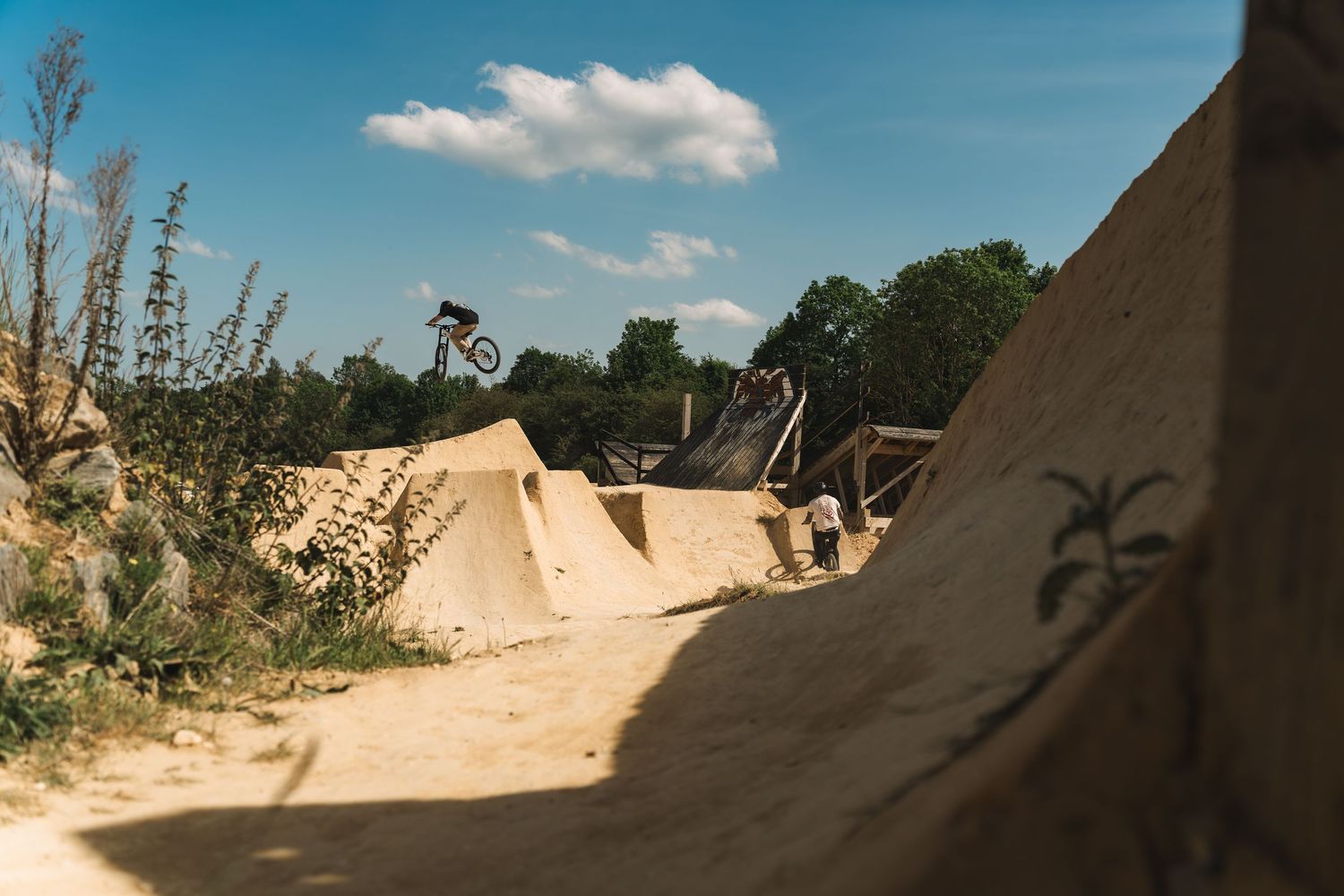 A BMX rider executes a trick between large sandy dirt jumps at an outdoor bike park.