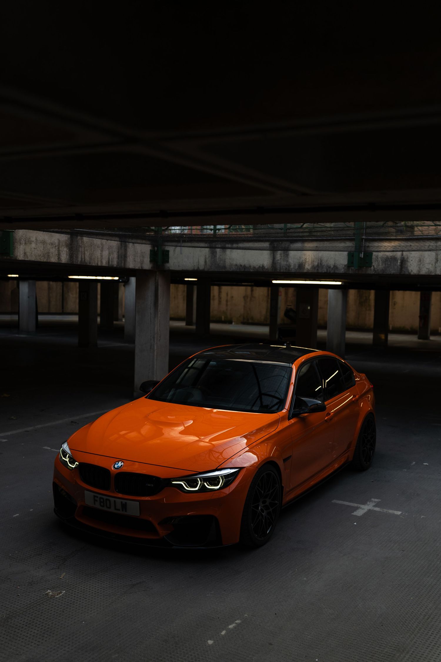 Vibrant orange BMW M3 sedan parked in a dark multi-story car park, showcasing its sleek design and angel eye headlights.