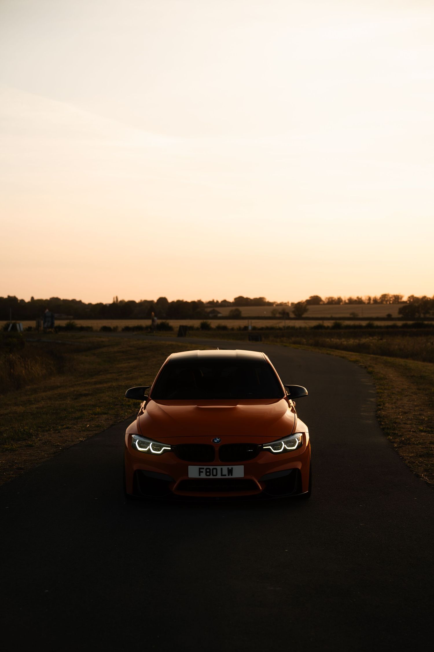 Red BMW M4 parked on a narrow road at sunset, with golden fields and a warm sky in the background.
