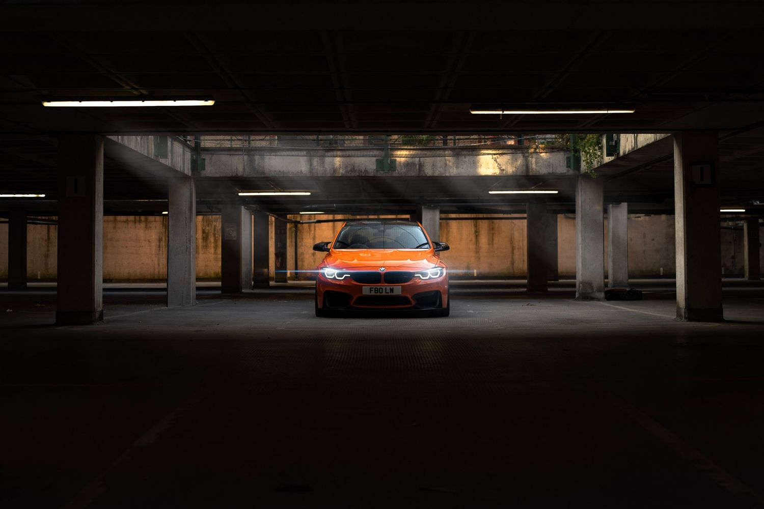 Orange Volkswagen sedan parked in a dark multi-story car park, illuminated by overhead fluorescent lights.