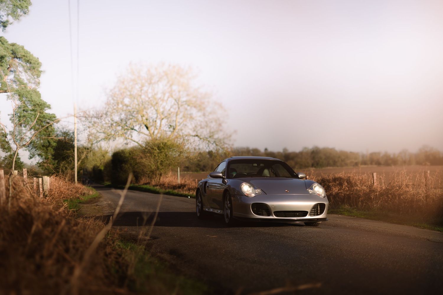 Silver Porsche 911 with headlights on driving along a misty rural road surrounded by autumn countryside.