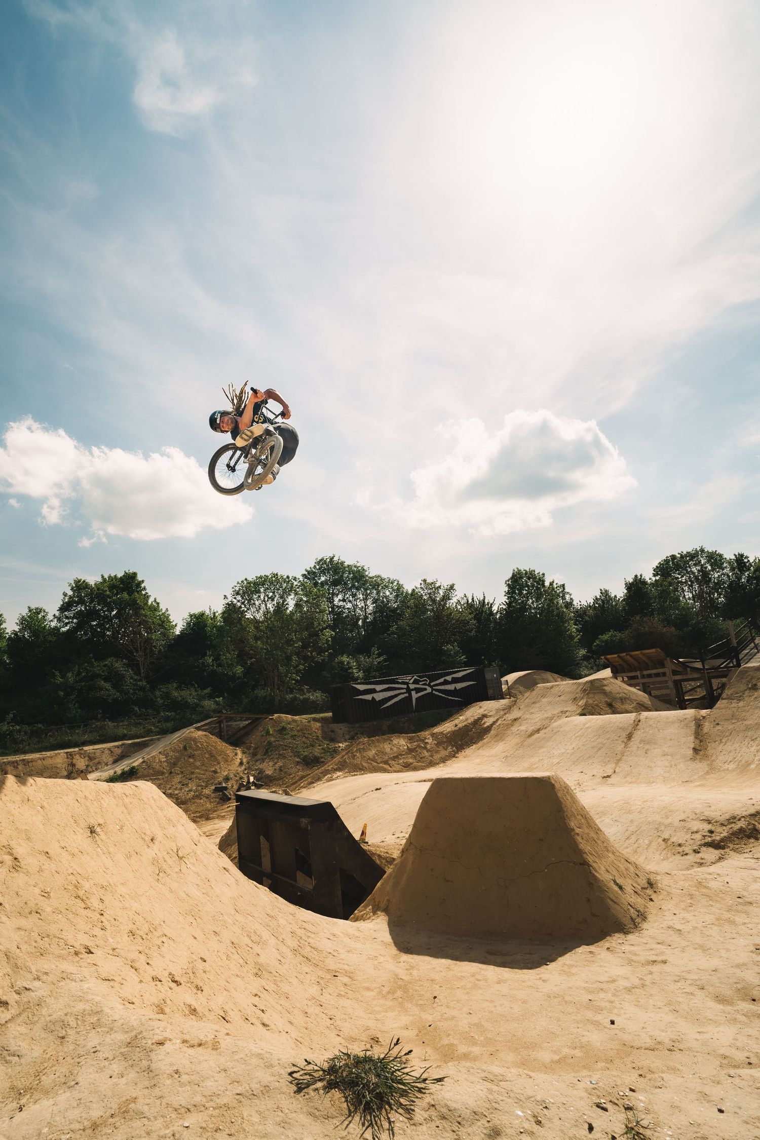 BMX rider performs a high aerial trick over dirt jumps at an outdoor bike park under a partly cloudy sky.