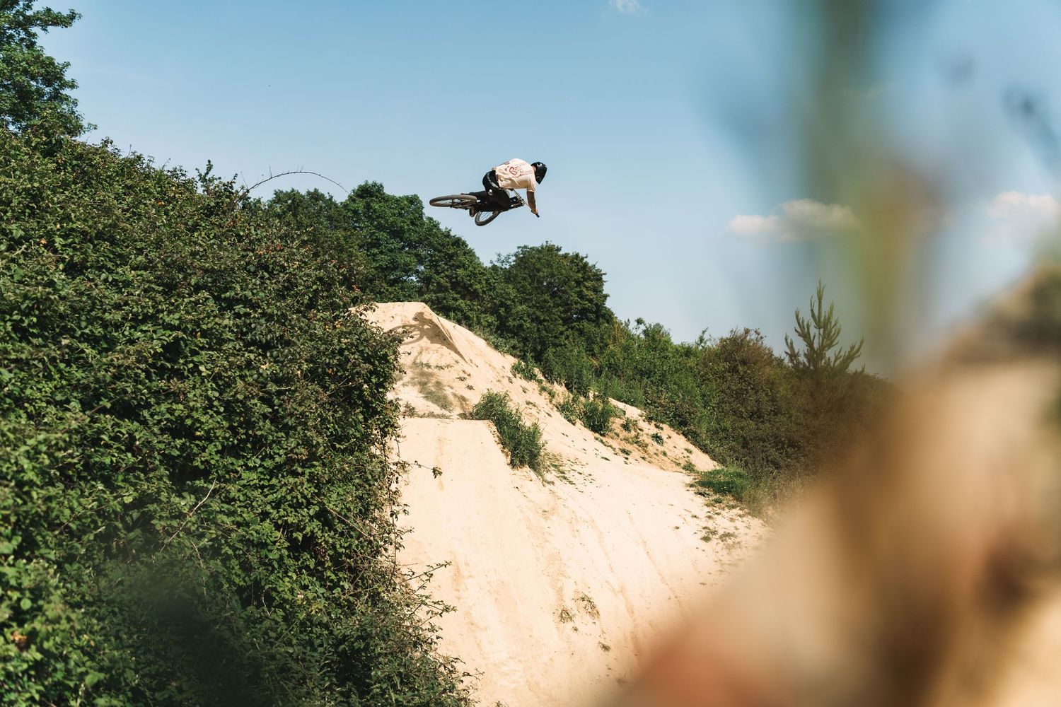 BMX rider launching off a sandy dirt jump hill surrounded by green trees under a clear blue sky.
