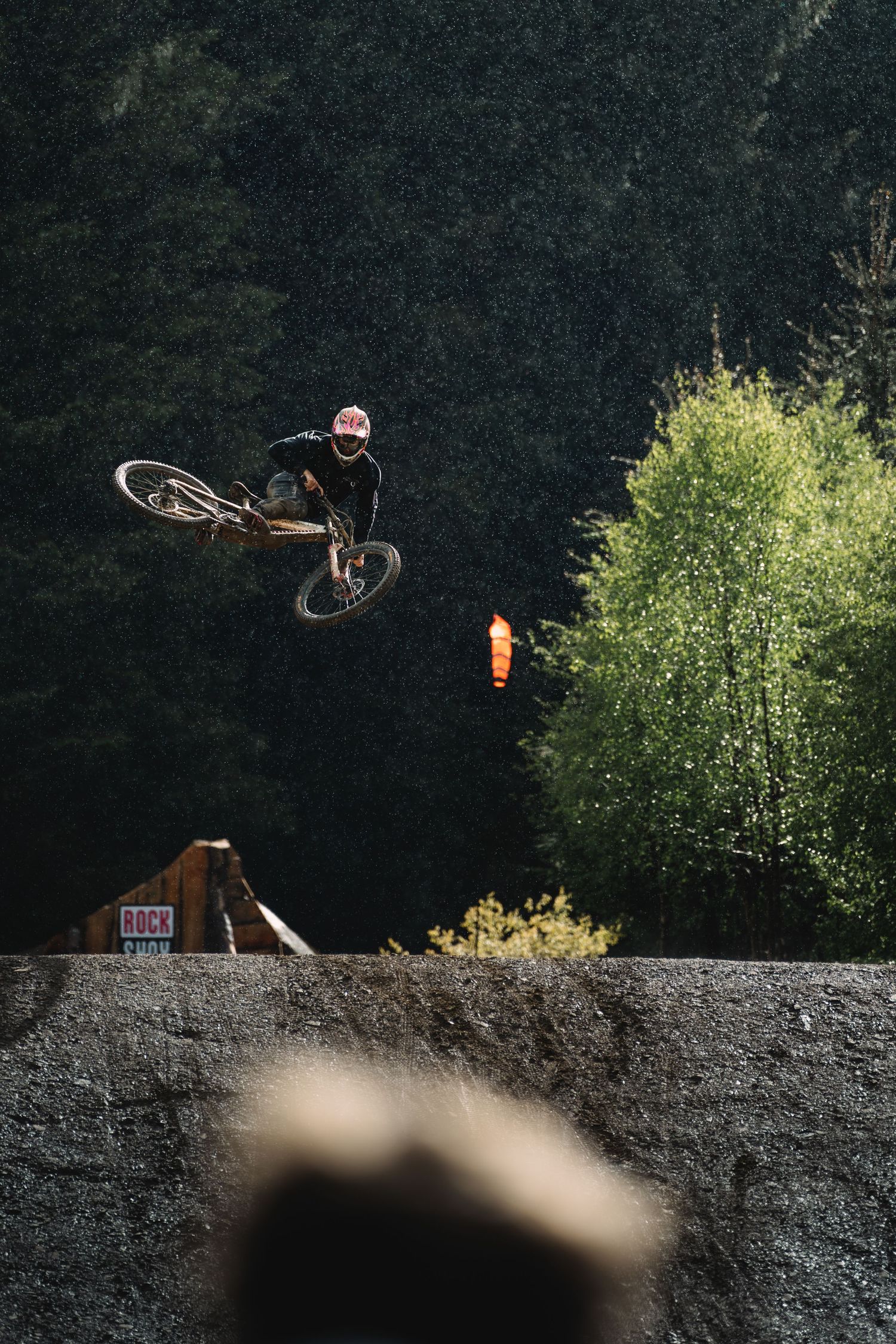 A mountain biker performs a dramatic aerial trick off a dirt ramp surrounded by lush green trees.