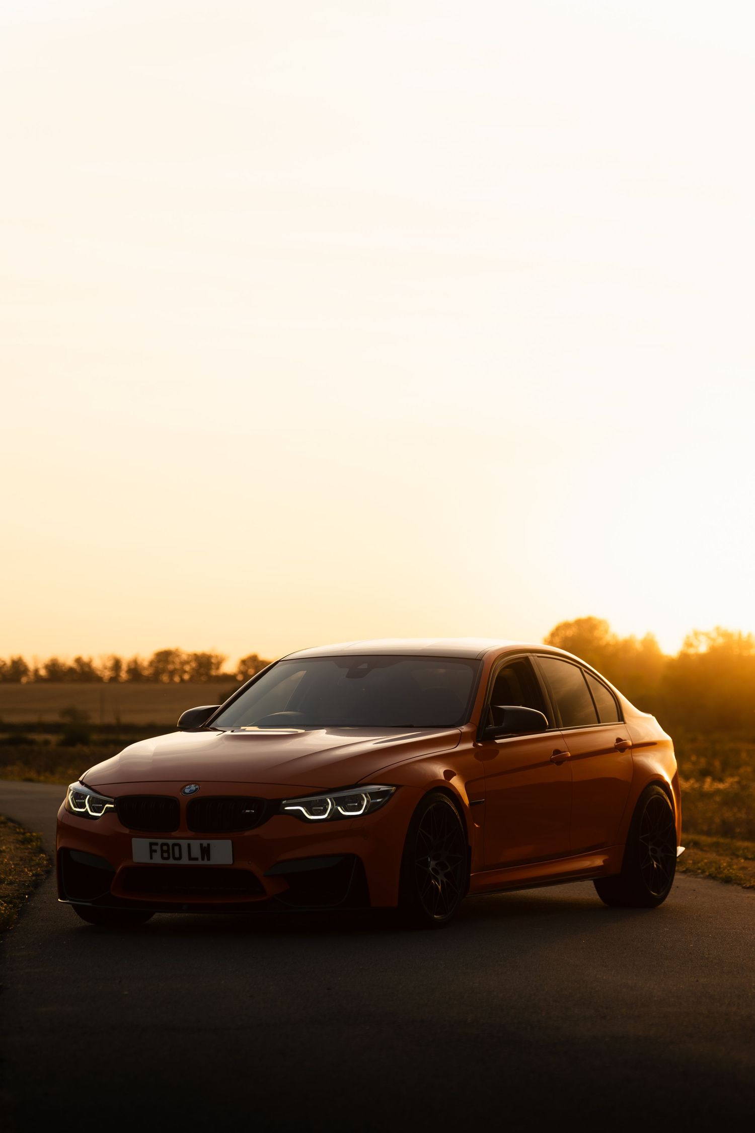 A stunning orange BMW M3 sedan parked on a road at golden hour sunset with warm glowing sky backdrop.