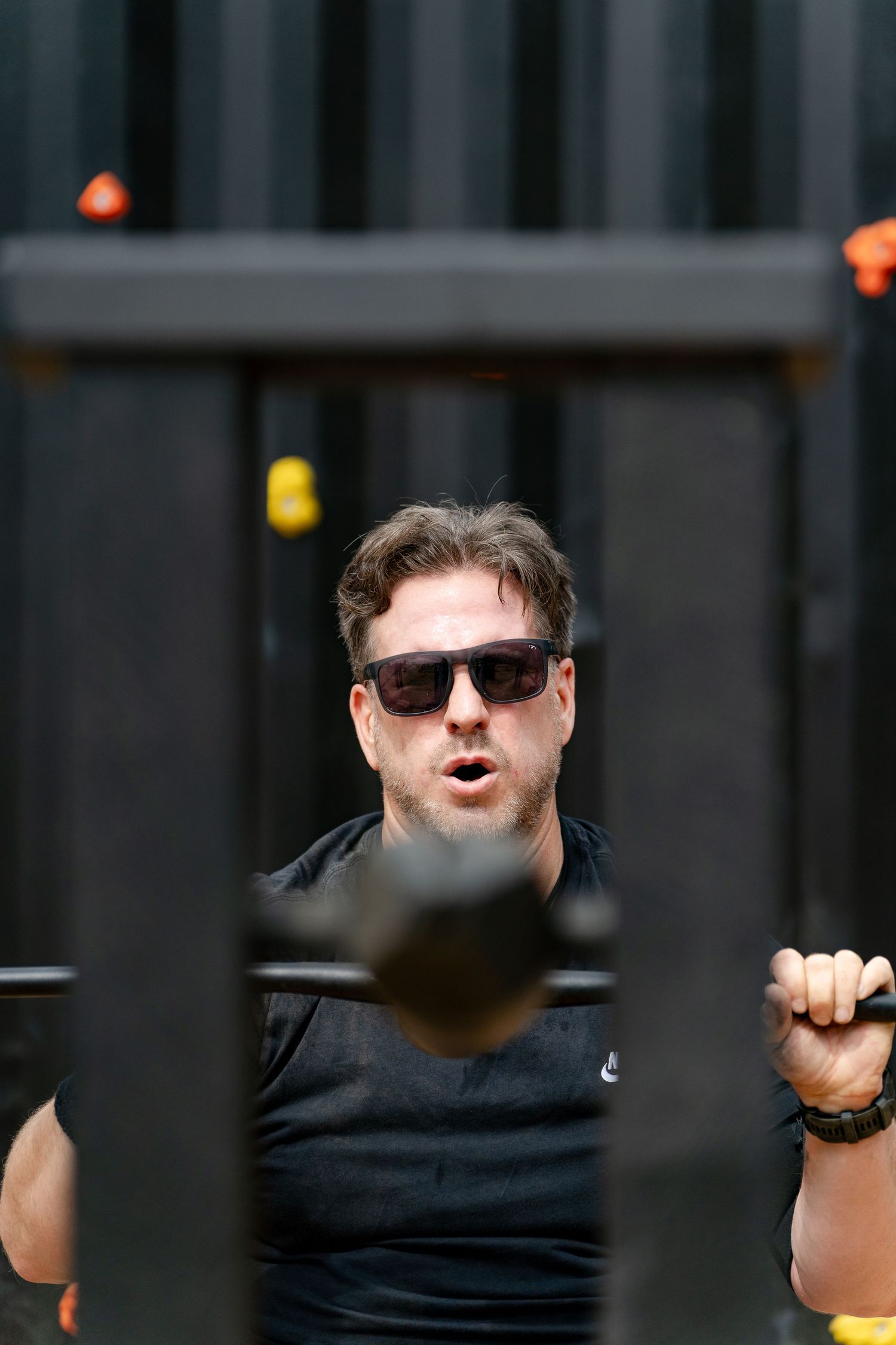 Man wearing sunglasses and black Nike shirt working out at an outdoor gym, gripping metal bars during exercise.