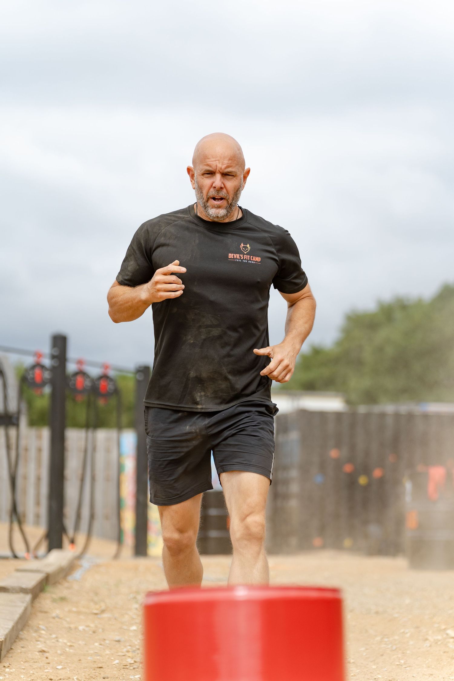 Bald man with beard running in muddy black t-shirt and shorts during outdoor obstacle course race.