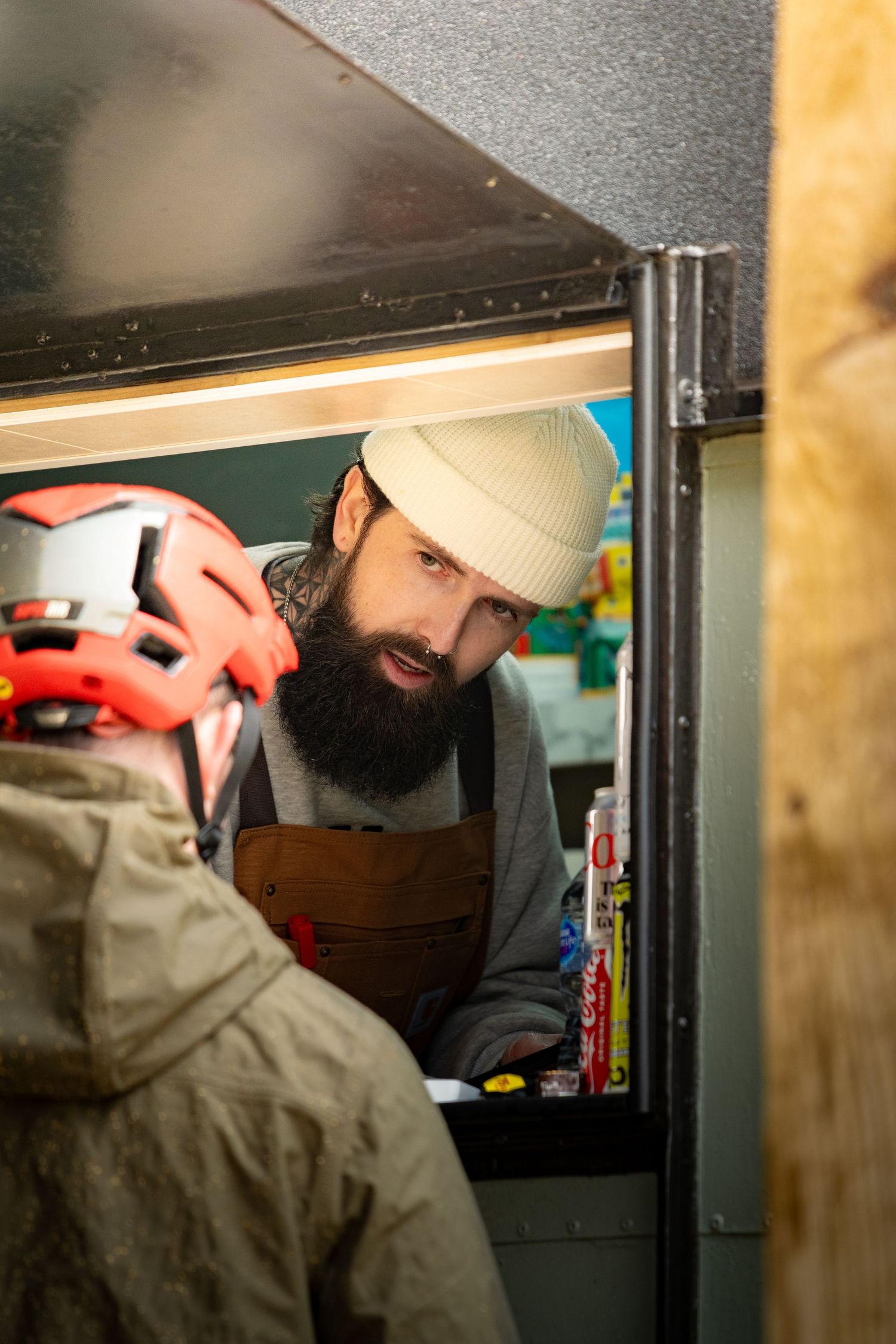Bearded barista in white beanie and brown apron serves customer through mobile coffee trailer window.