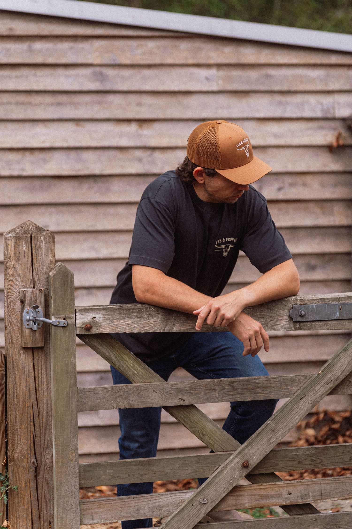 Man in brown cap and dark t-shirt leaning on a rustic wooden farm gate outdoors.
