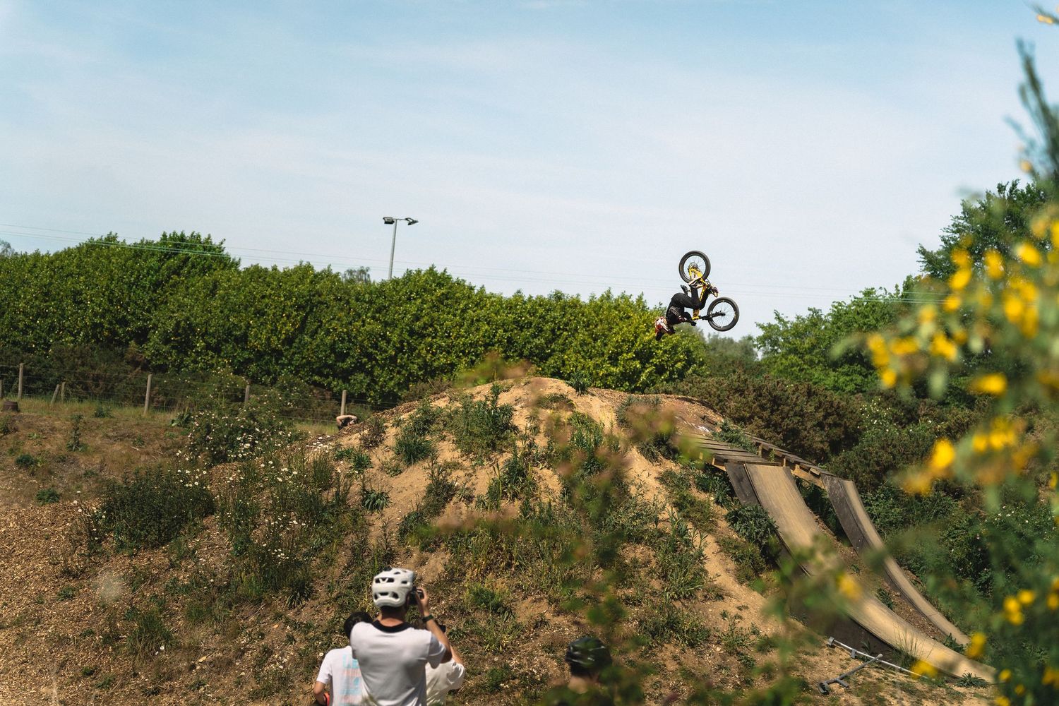 BMX rider performing a high jump trick over a dirt mound at an outdoor bike park with spectator watching.