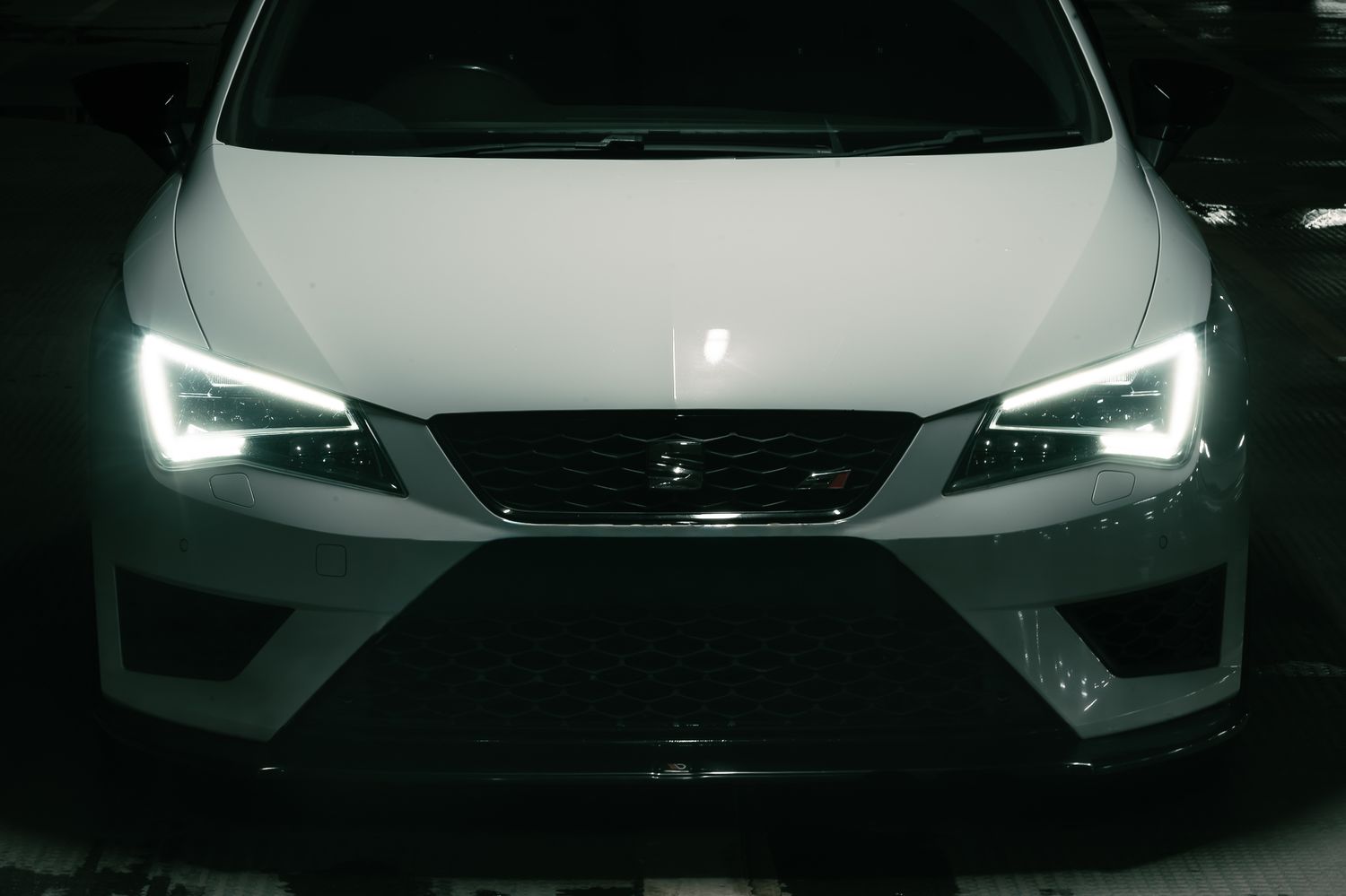 Front view of a white SEAT Leon Cupra with bright LED headlights glowing in a dark parking garage.