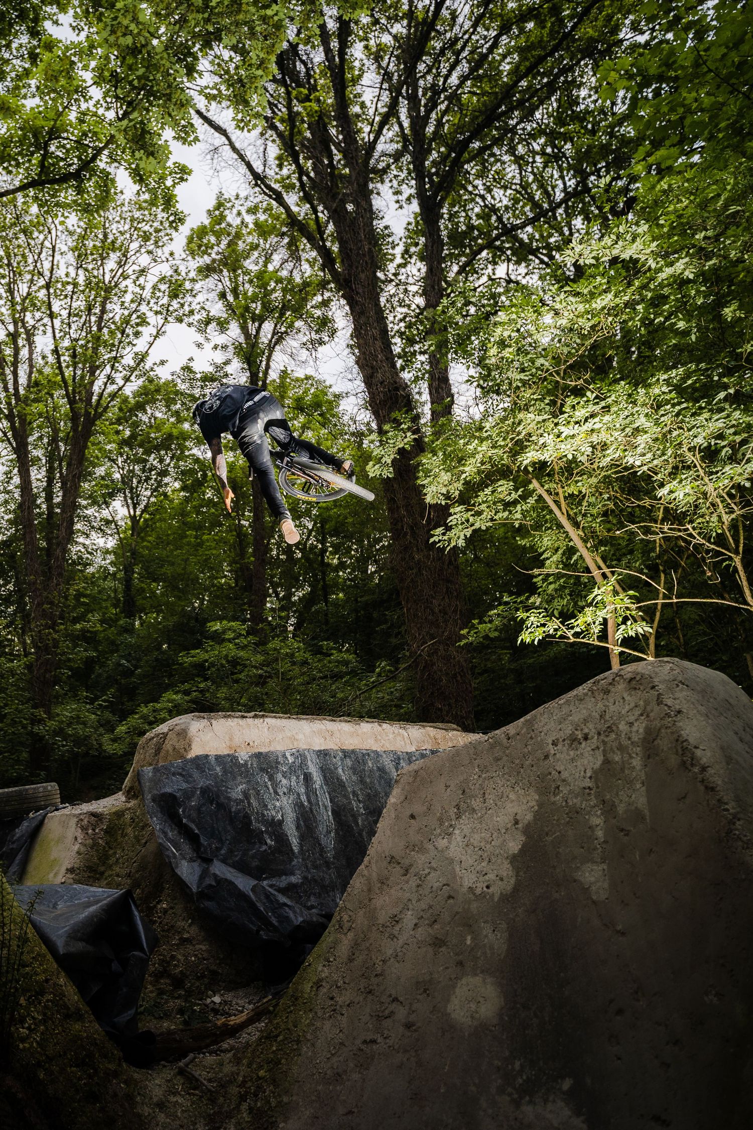 Skateboarder performing aerial trick over large rocks in a lush green forest setting.