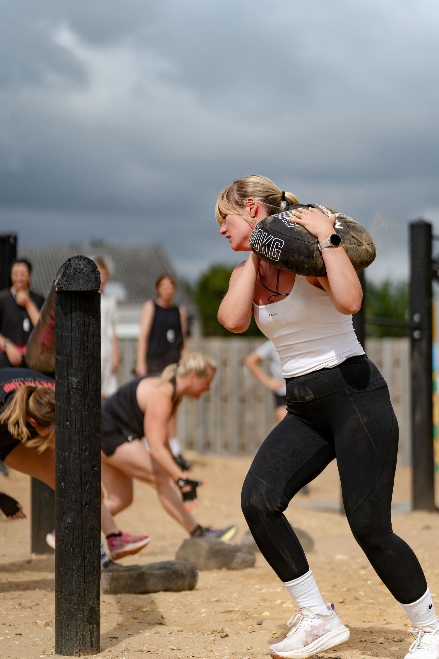 Woman carrying heavy sandbag on shoulder during outdoor fitness competition on sandy ground with cloudy sky.